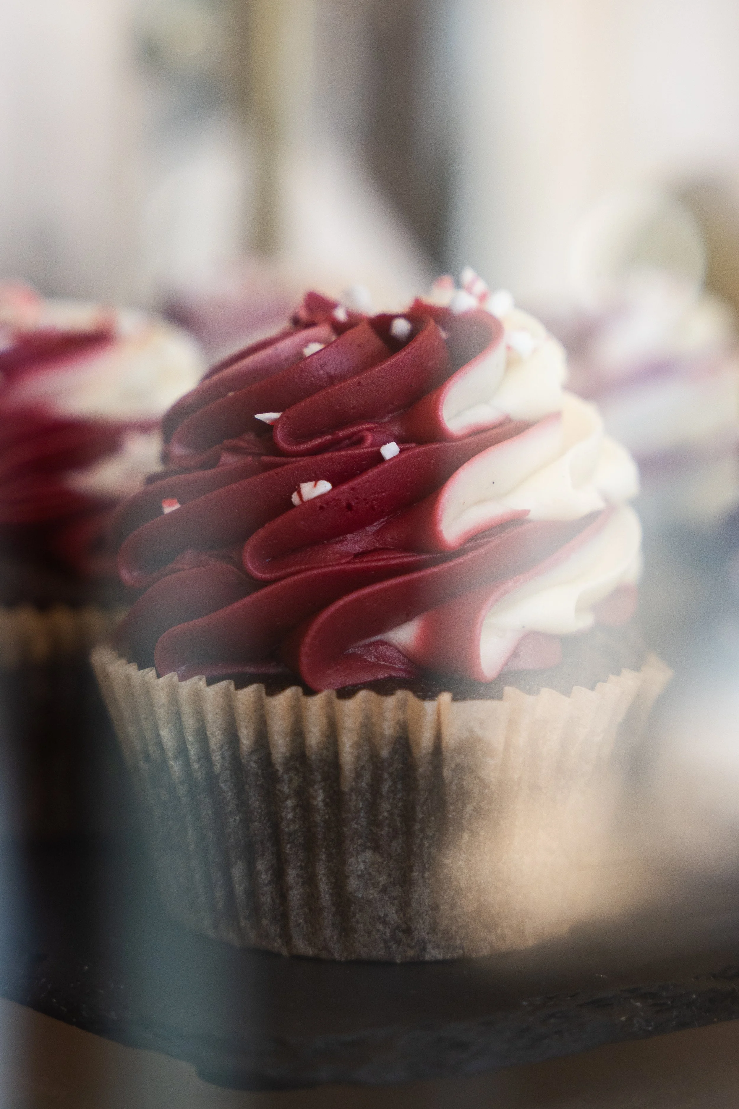 A cupcake with red and white swirled frosting topped with small white sprinkles, inside a paper cup.