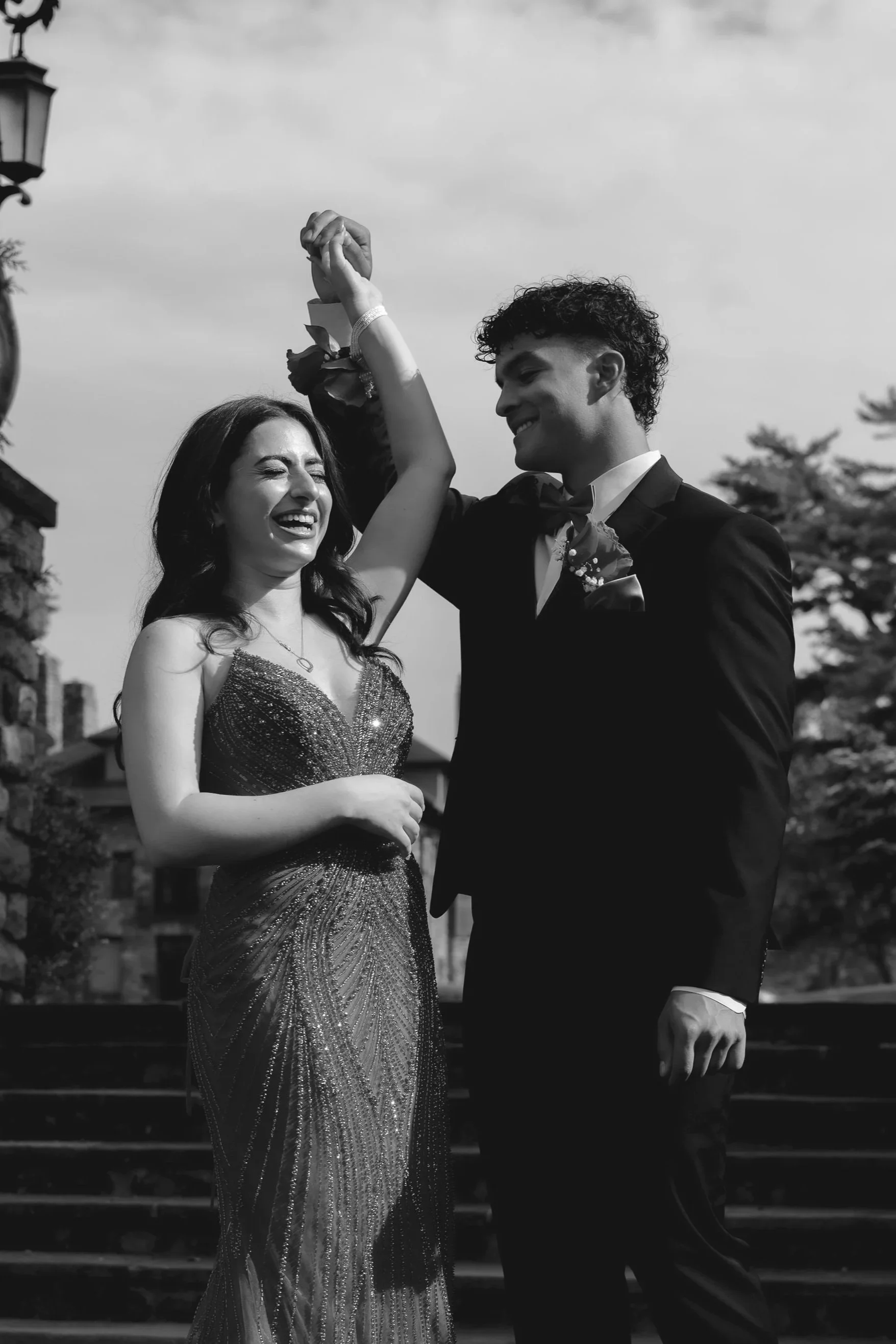 A couple in formal attire dancing outdoors, smiling and holding hands, with trees and a building in the background.