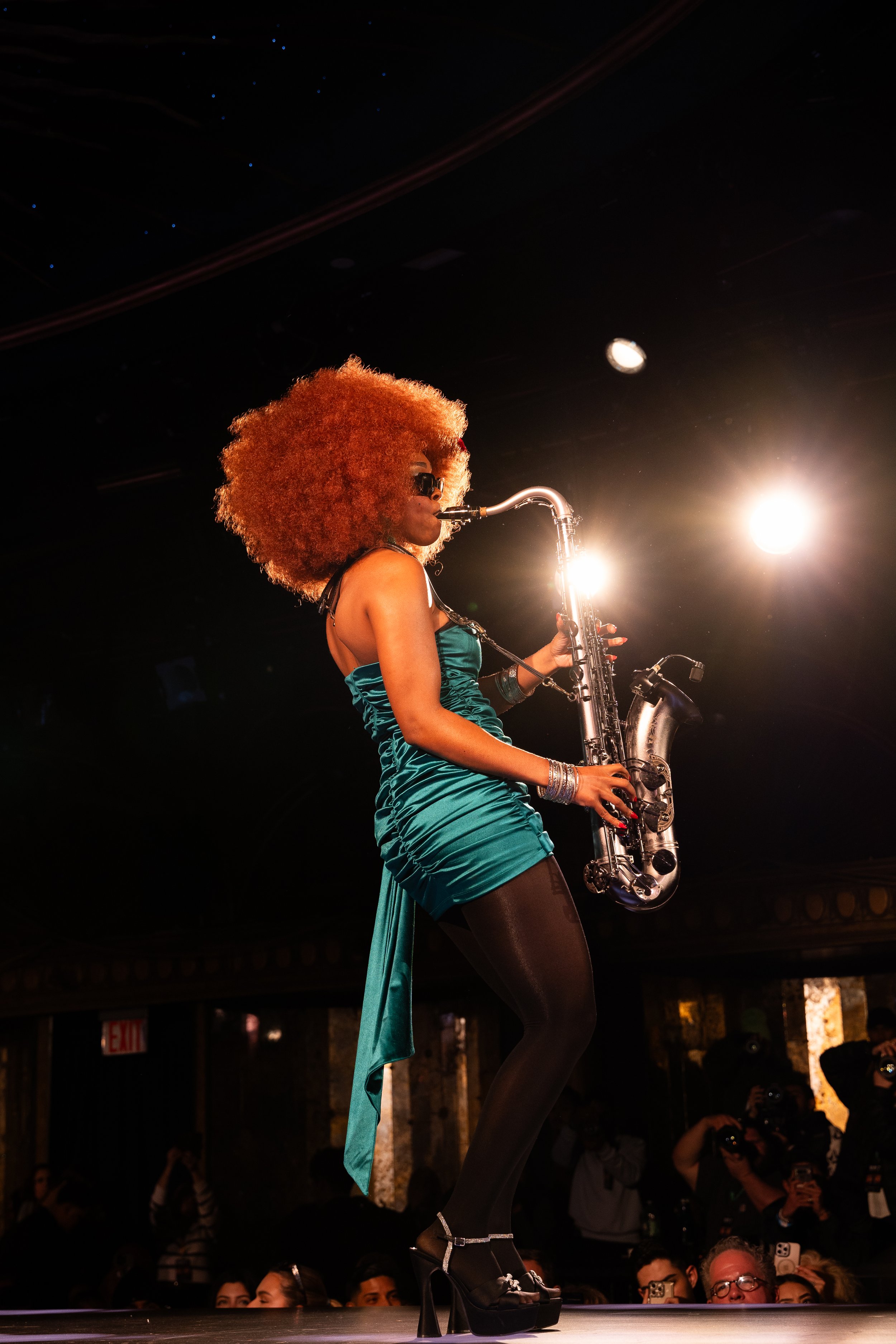 A woman with large curly red hair wearing sunglasses, a teal dress, and high heels is playing a saxophone on stage during a performance. She is elevated on a platform with an audience watching her.