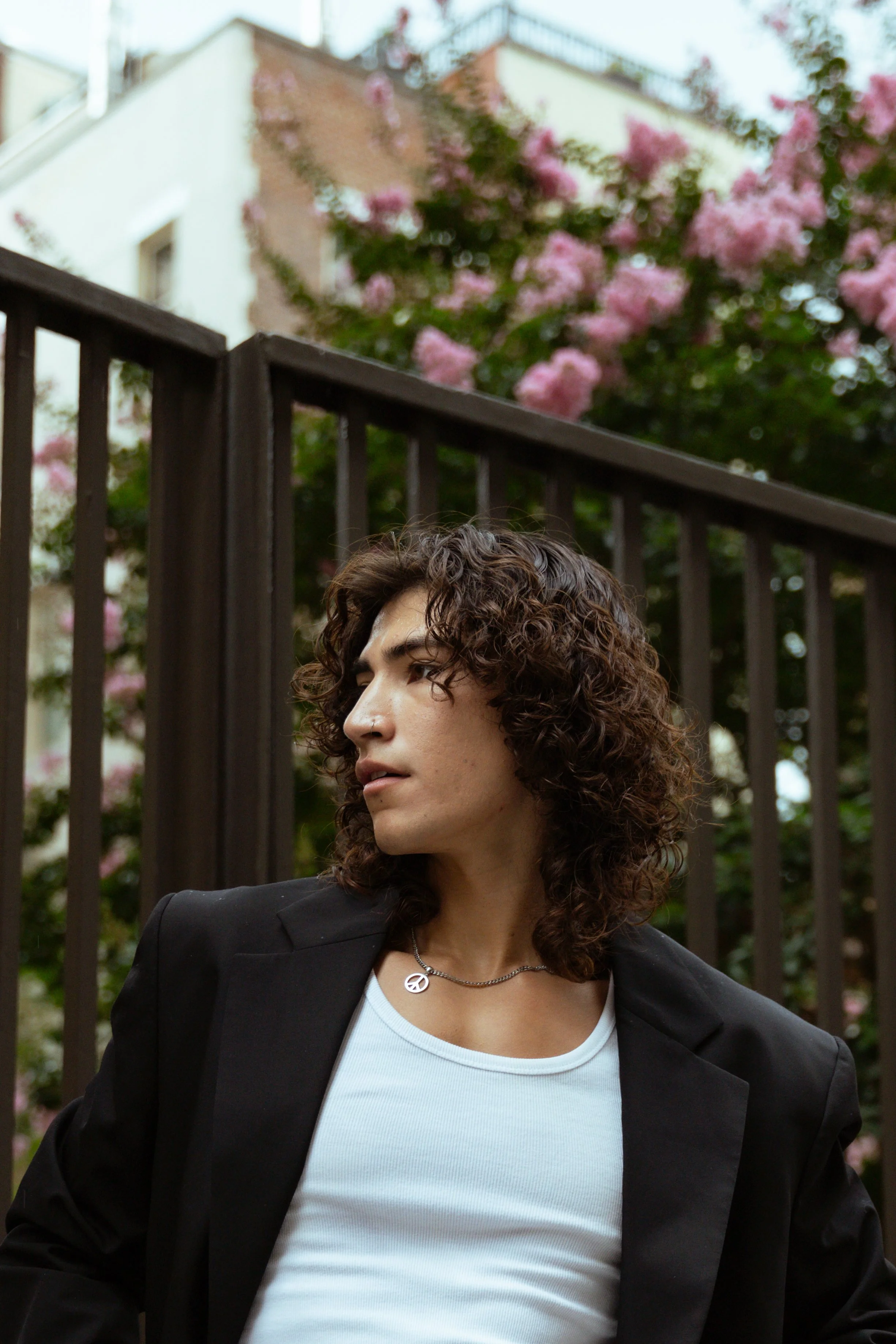 A young woman with curly brown hair wearing a black blazer, light blue T-shirt, a peace sign necklace, standing outdoors near a black metal fence with pink flowering trees and buildings in the background.