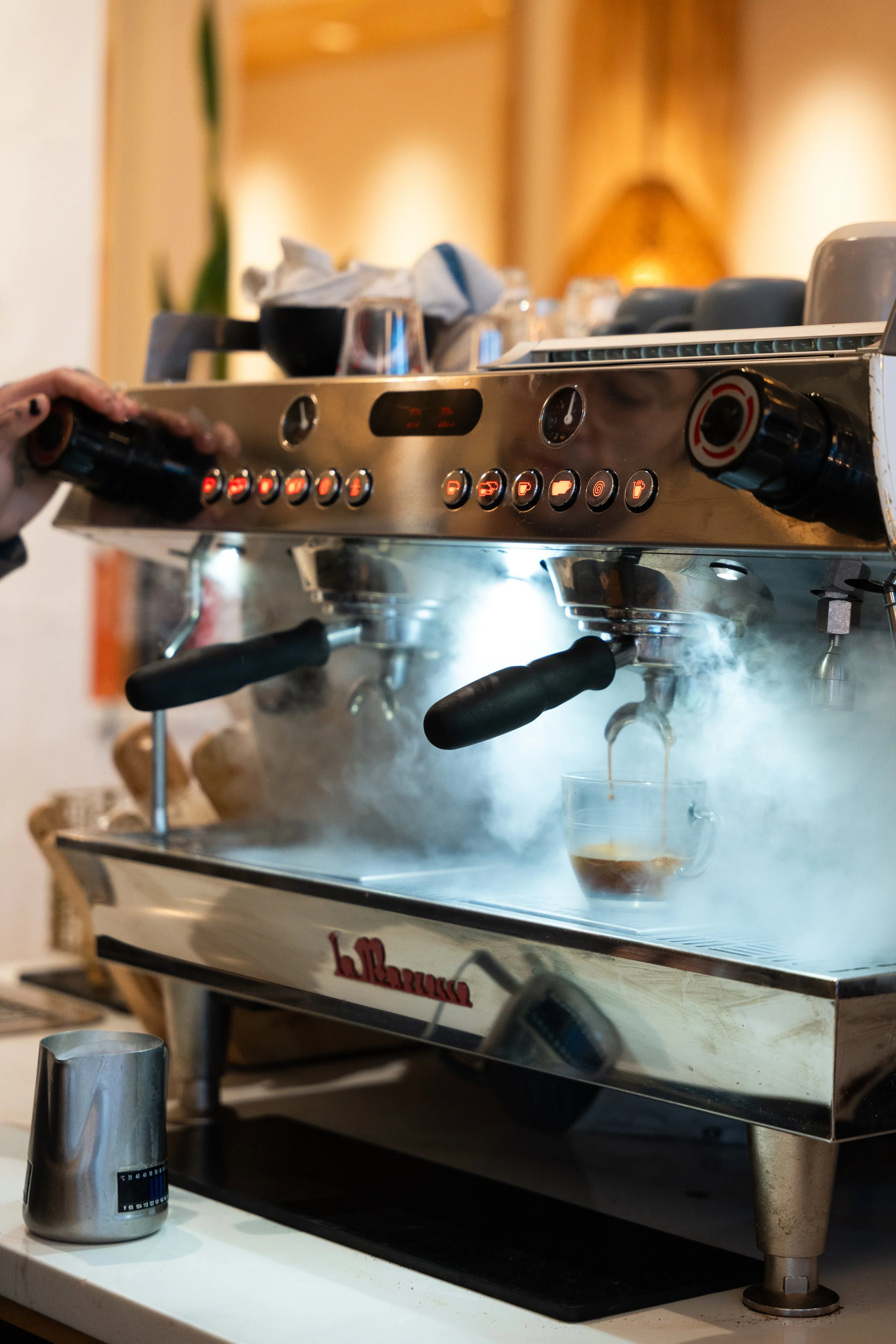 A barista is preparing an espresso shot in a commercial coffee machine with steam rising, with cups and glasses on a counter in the background.