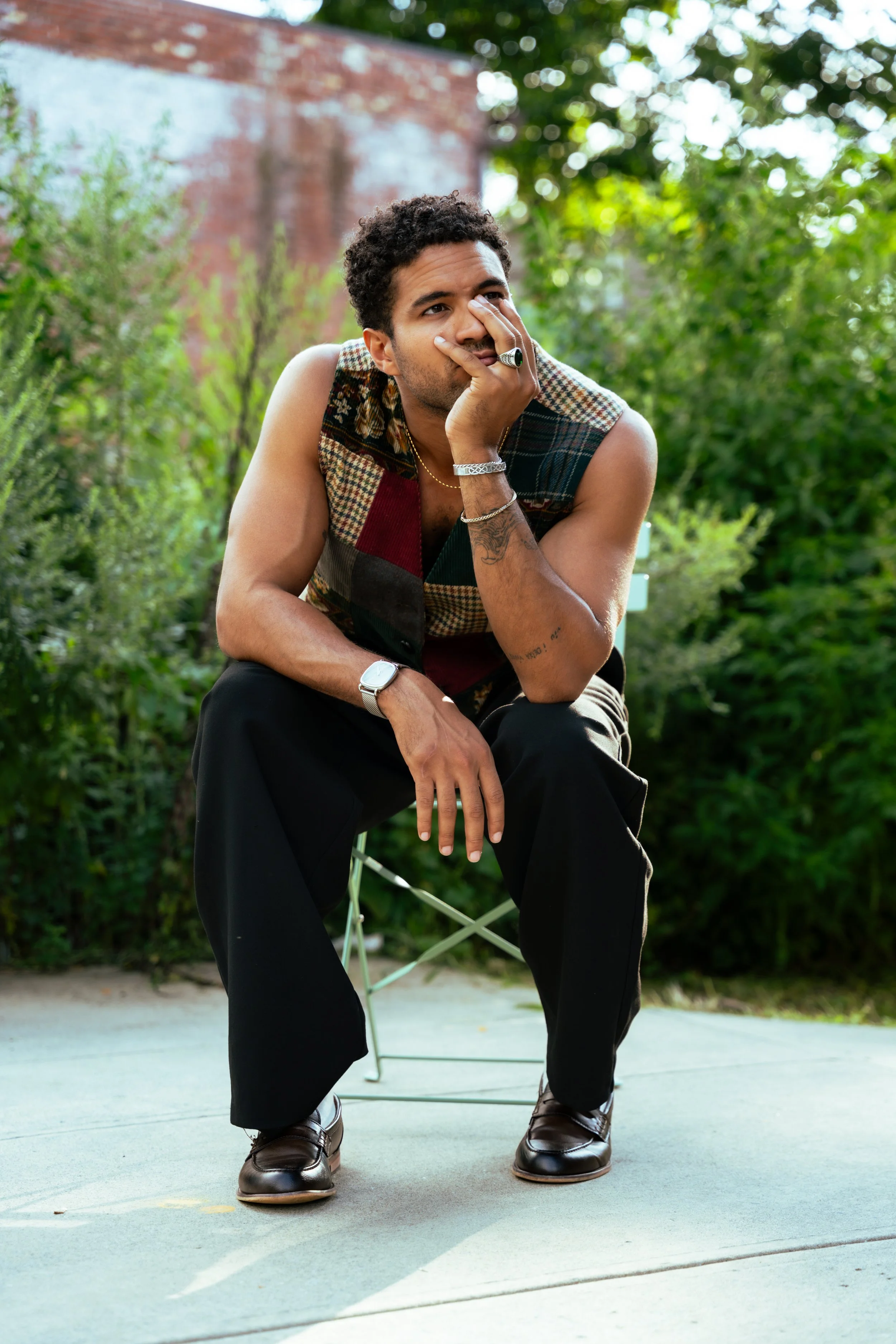 A young man with curly hair, beard, in a sleeveless shirt, sitting on a portable chair outdoors on a gray concrete surface, with an outdoor background of green bushes and trees, and a brick building in the distance.