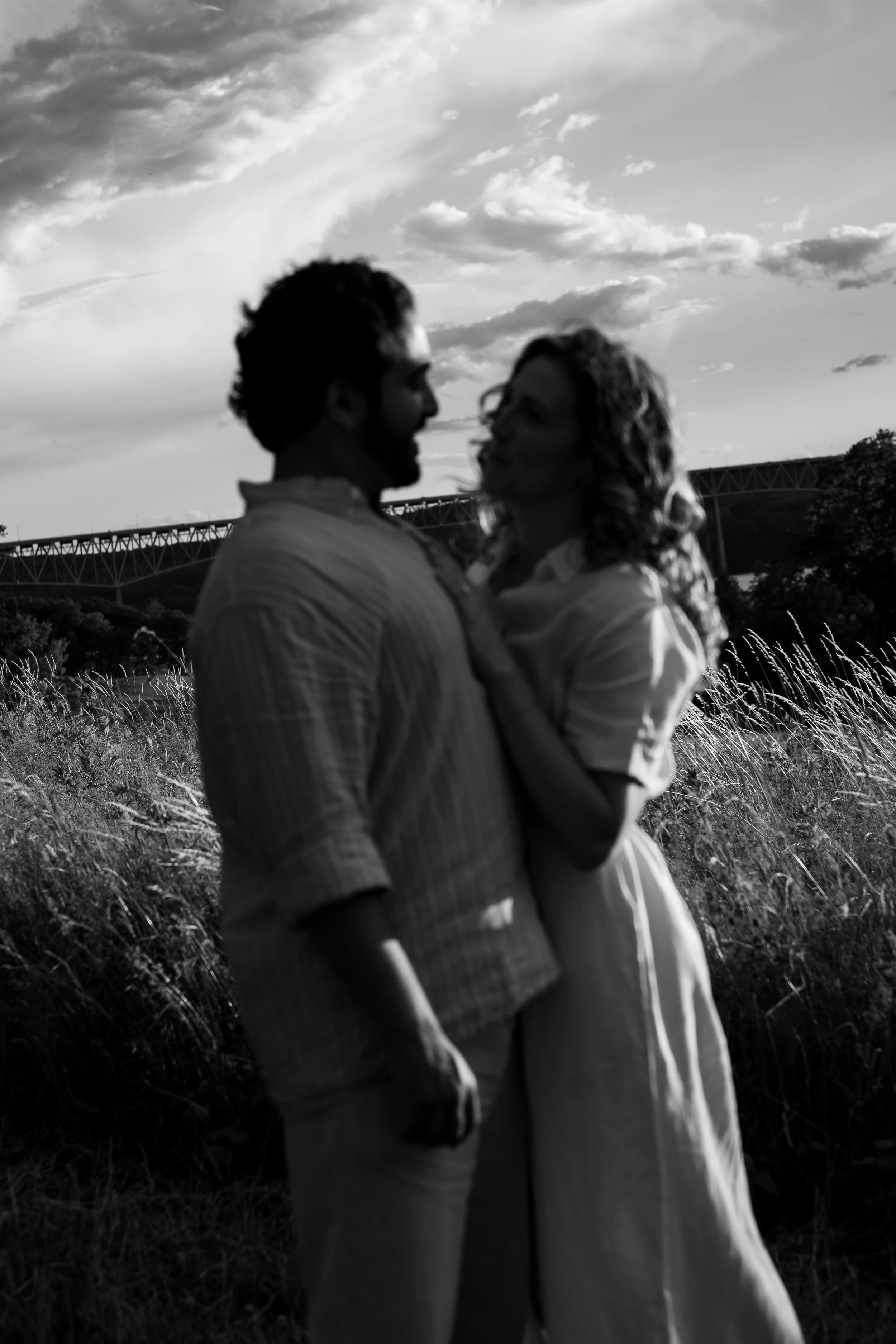A black and white photo of a couple standing outdoors, facing each other, with the man on the left and the woman on the right. They are close and appear to be about to kiss or share an intimate moment. The background shows a sky with clouds, a bridge
