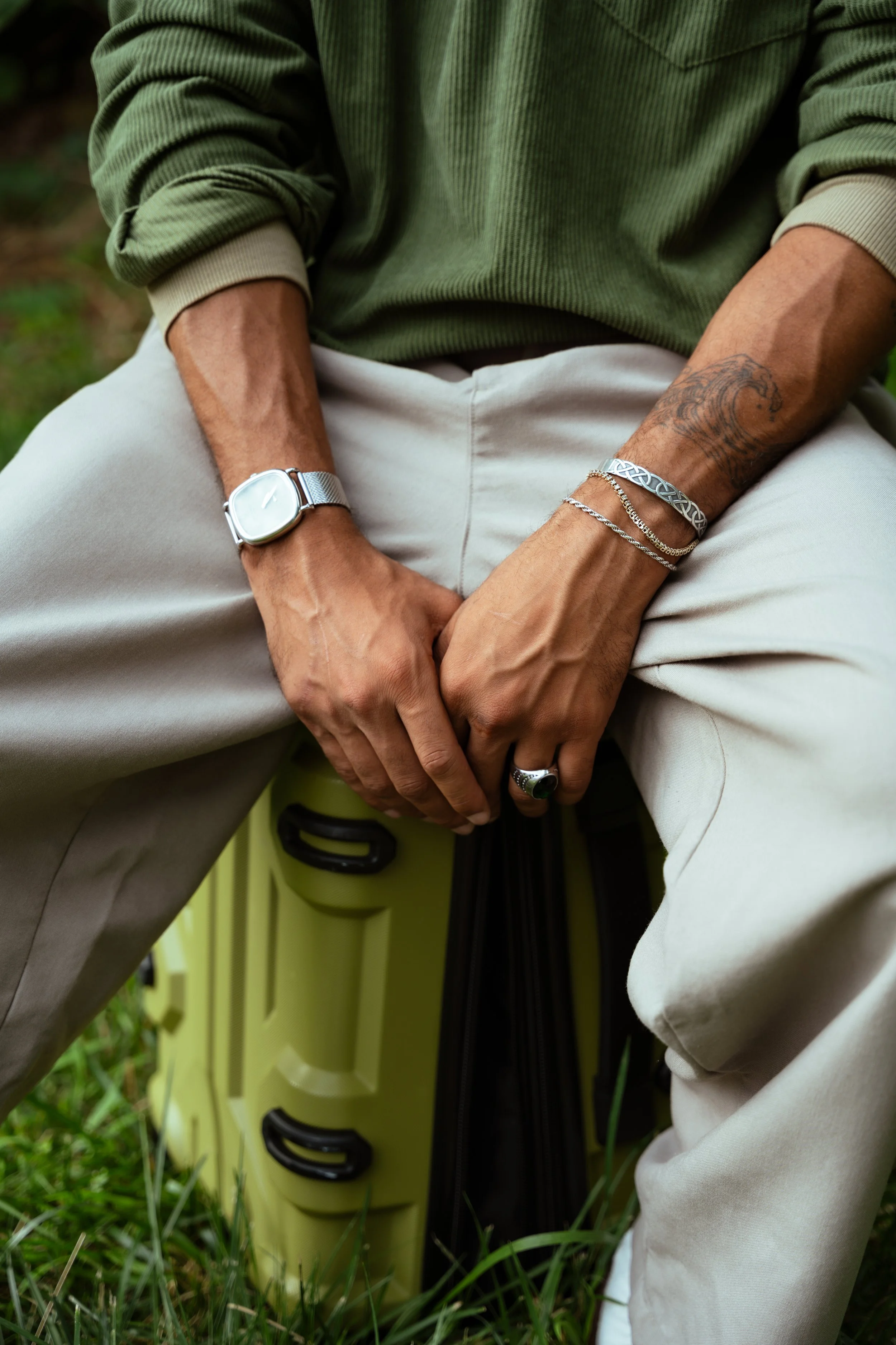 A person sitting outdoors wearing a green long-sleeve shirt and beige pants, showing a watch on the left wrist, multiple bracelets on the right wrist, and a ring on the right hand. The person has a tattoo on the right forearm and is sitting next to a
