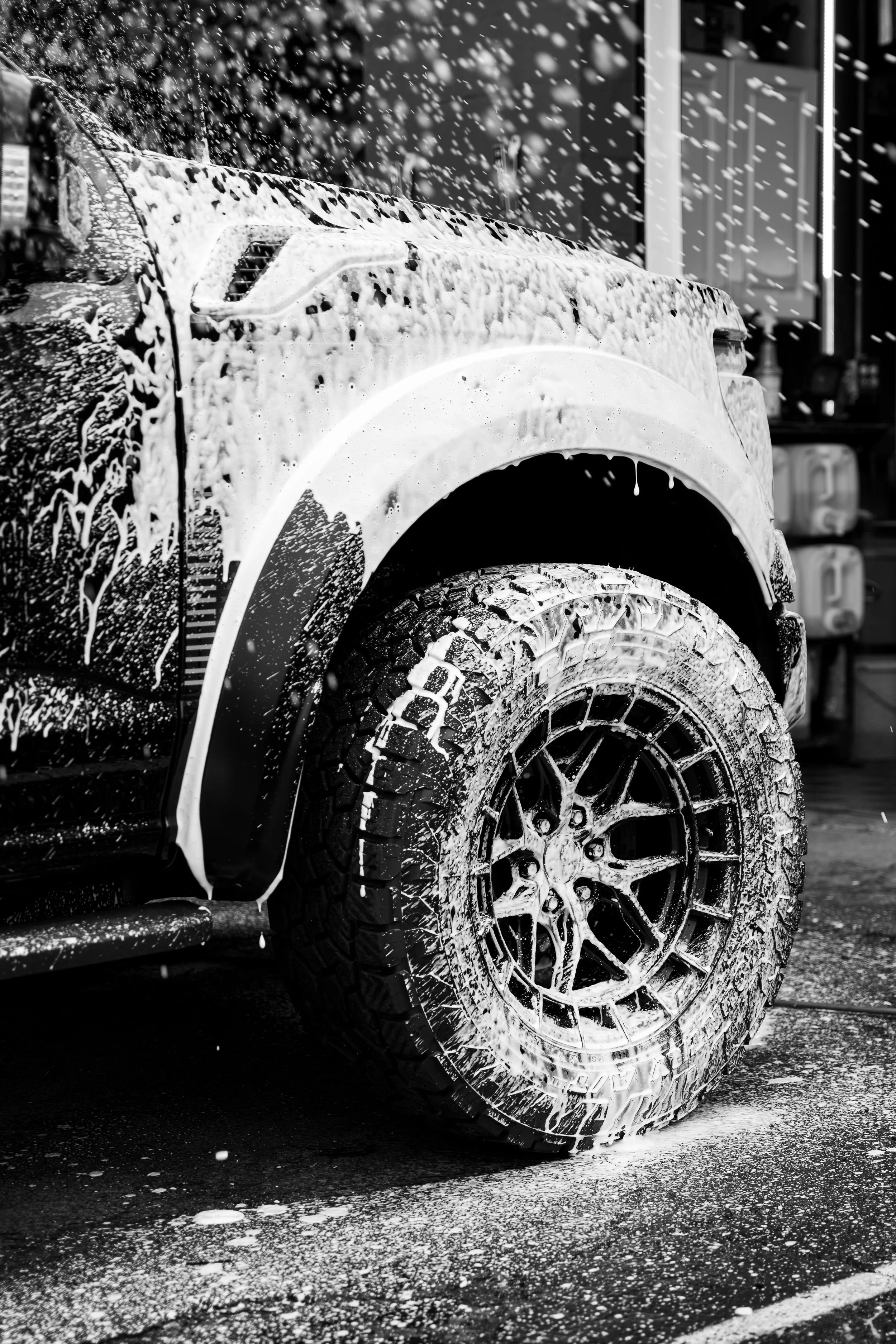 Close-up of a vehicle's front tire and wheel being washed with foam and water, with soap suds on the tire and ground.