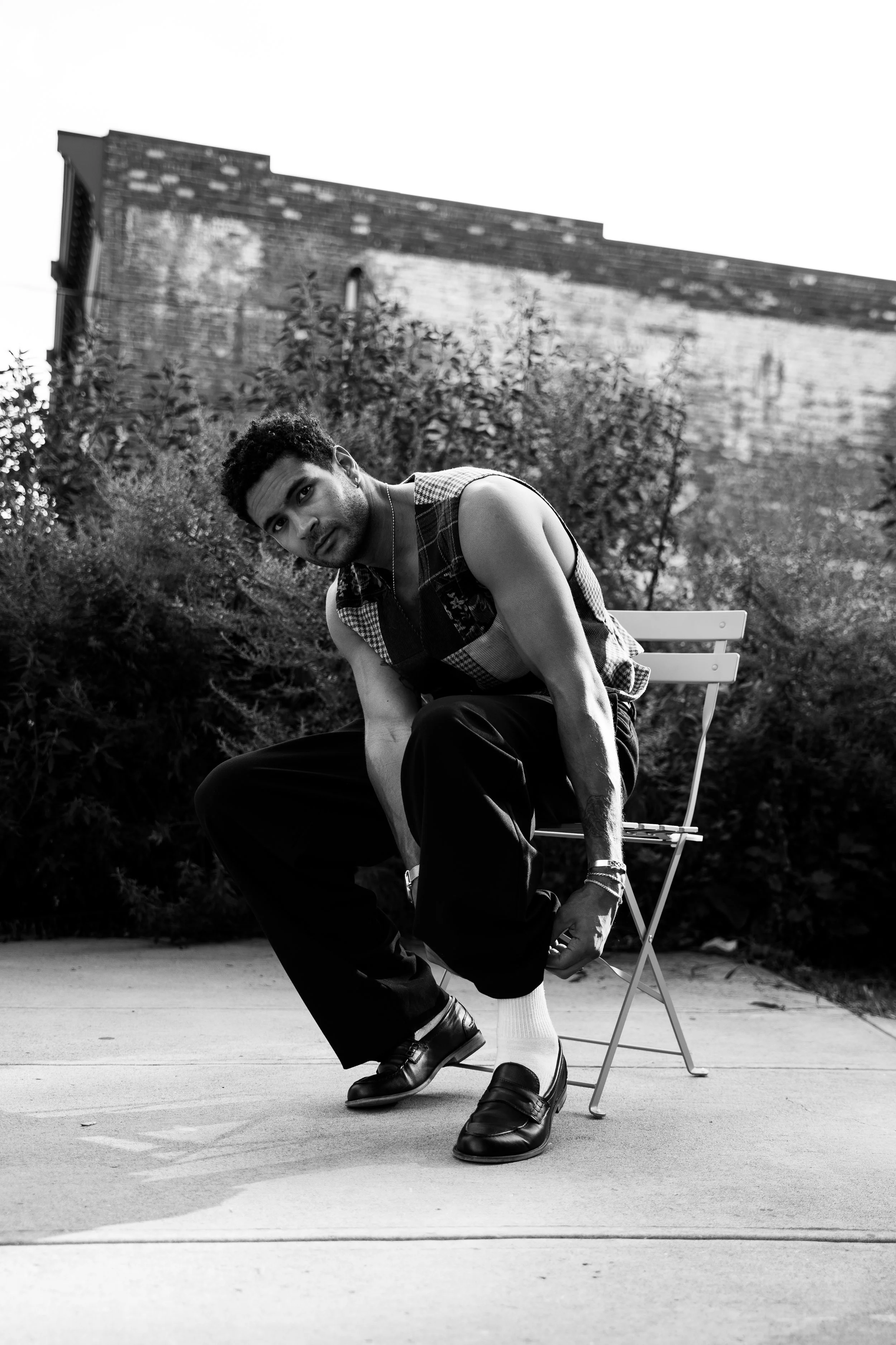 A young man with curly hair and a sleeveless shirt sitting on a chair outdoors, adjusting his shoe, with a brick building and greenery in the background, in black and white.