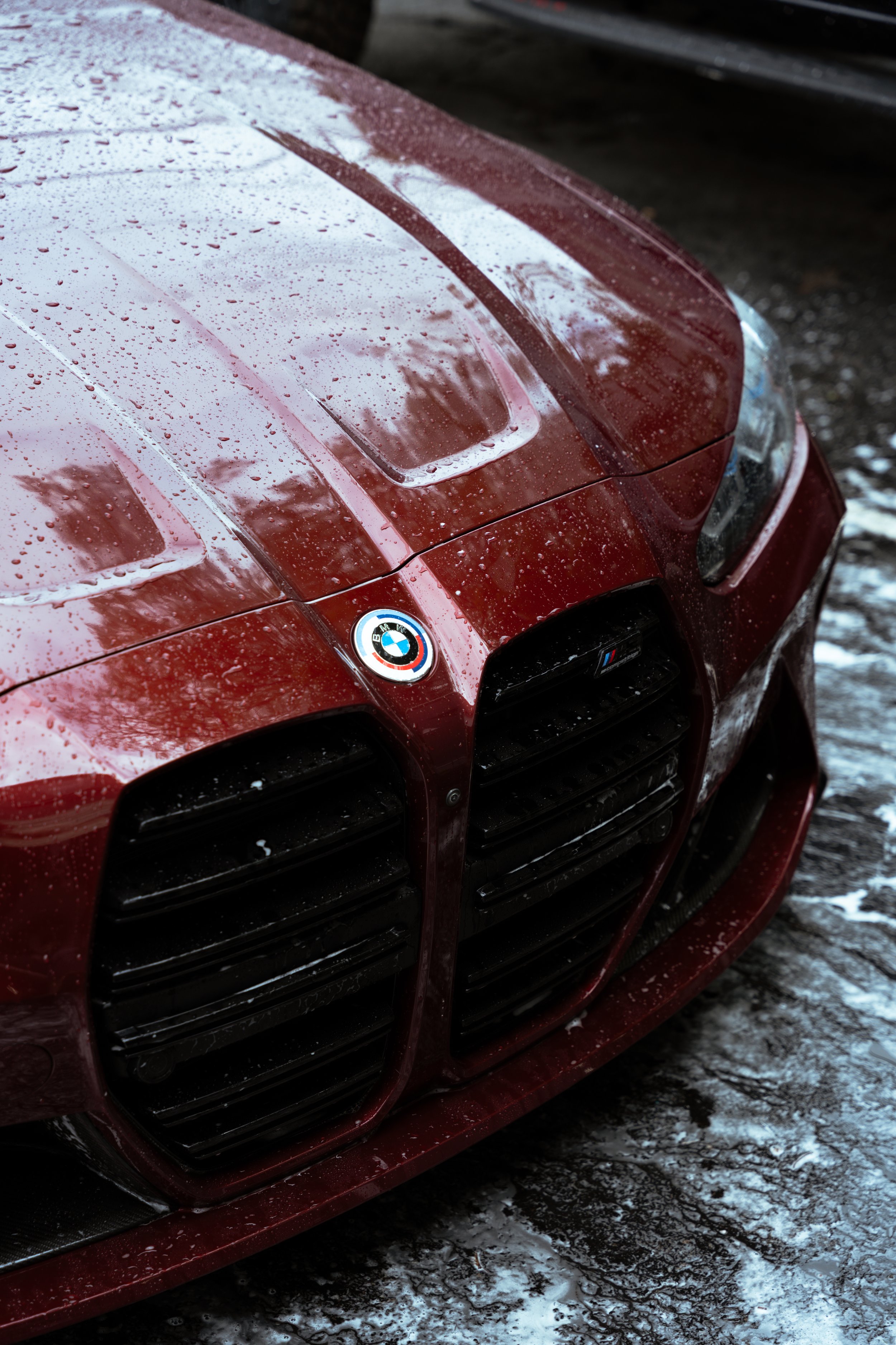 Close-up of the front of a red BMW car with water droplets on its surface, parked on a wet, snowy ground.