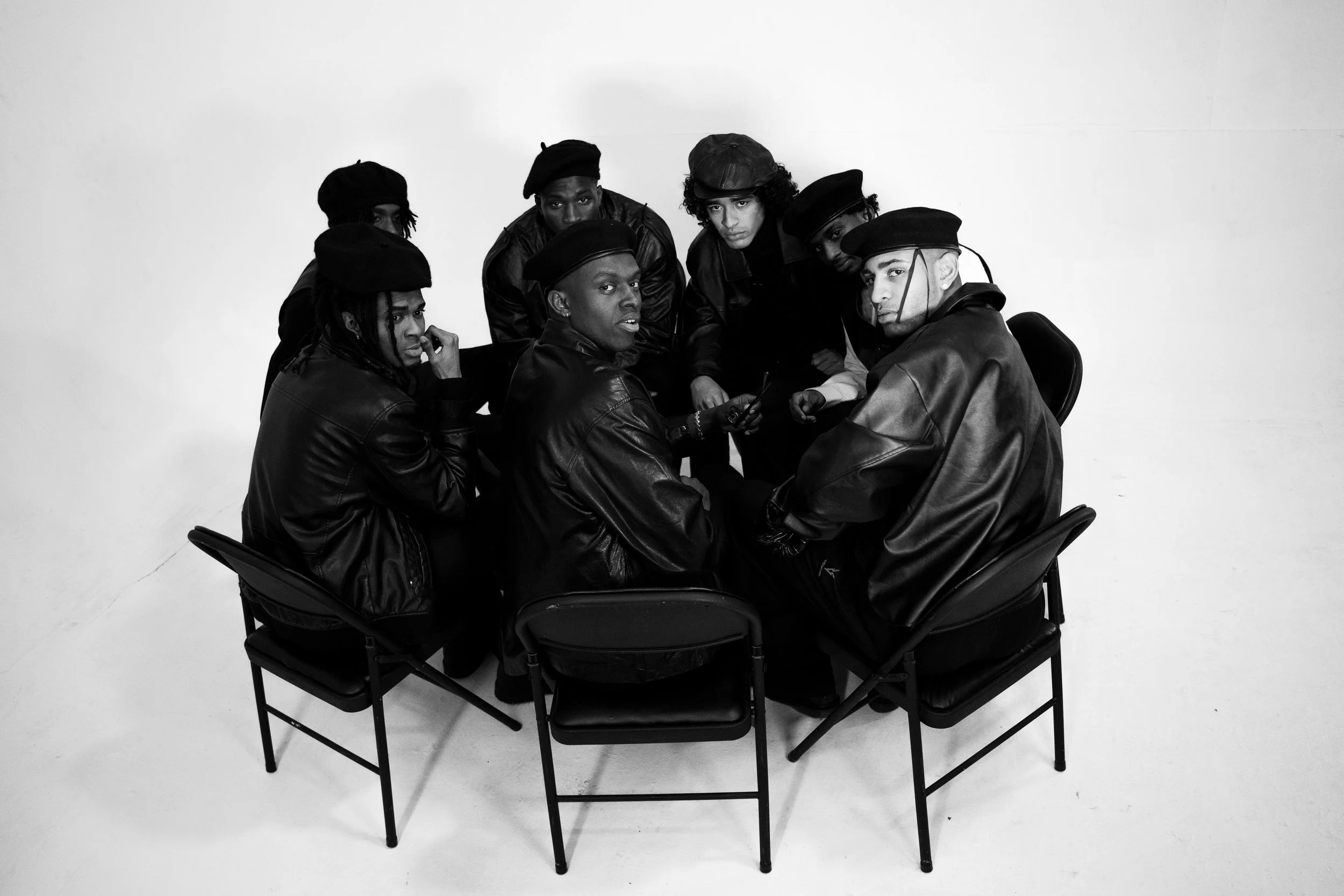 A group of seven men sitting around a table in a photo studio with a white background. They are dressed in dark leather jackets and hats, some are wearing berets and others are wearing caps. The men have serious expressions and are looking towards th