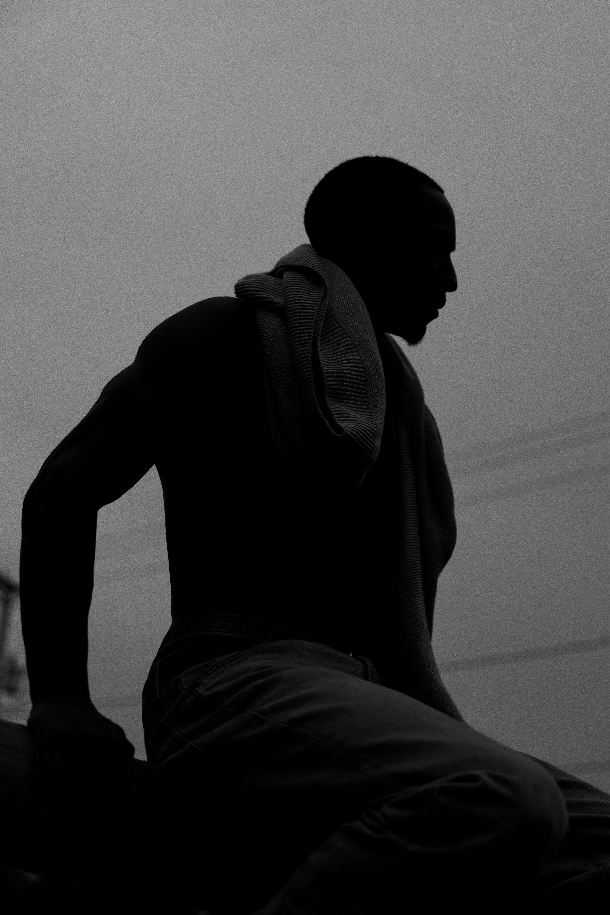 Silhouette of a man with a shaved head, sitting outdoors during dusk or dawn, with a scarf around his neck and dark clothing, against a gray sky.