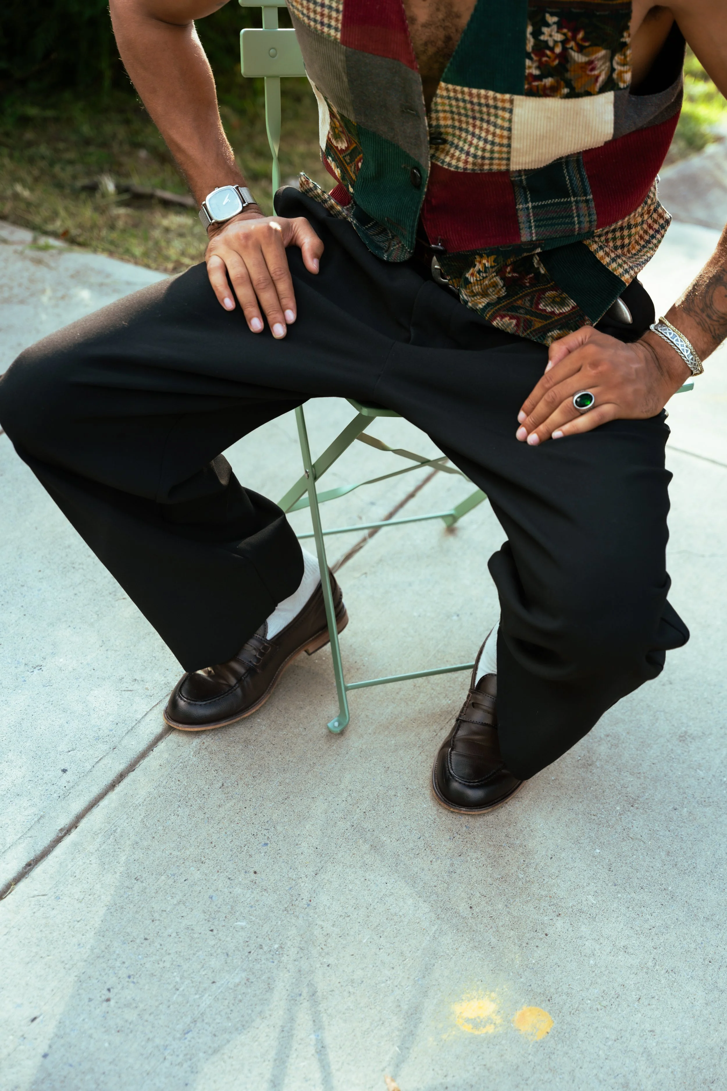 A man sitting outdoors on a green folding chair with his left hand resting on his thigh and his right hand on his knee. He is wearing a colorful patchwork vest, black pants, brown leather shoes, a silver wristwatch, a silver ring with a green stone, 