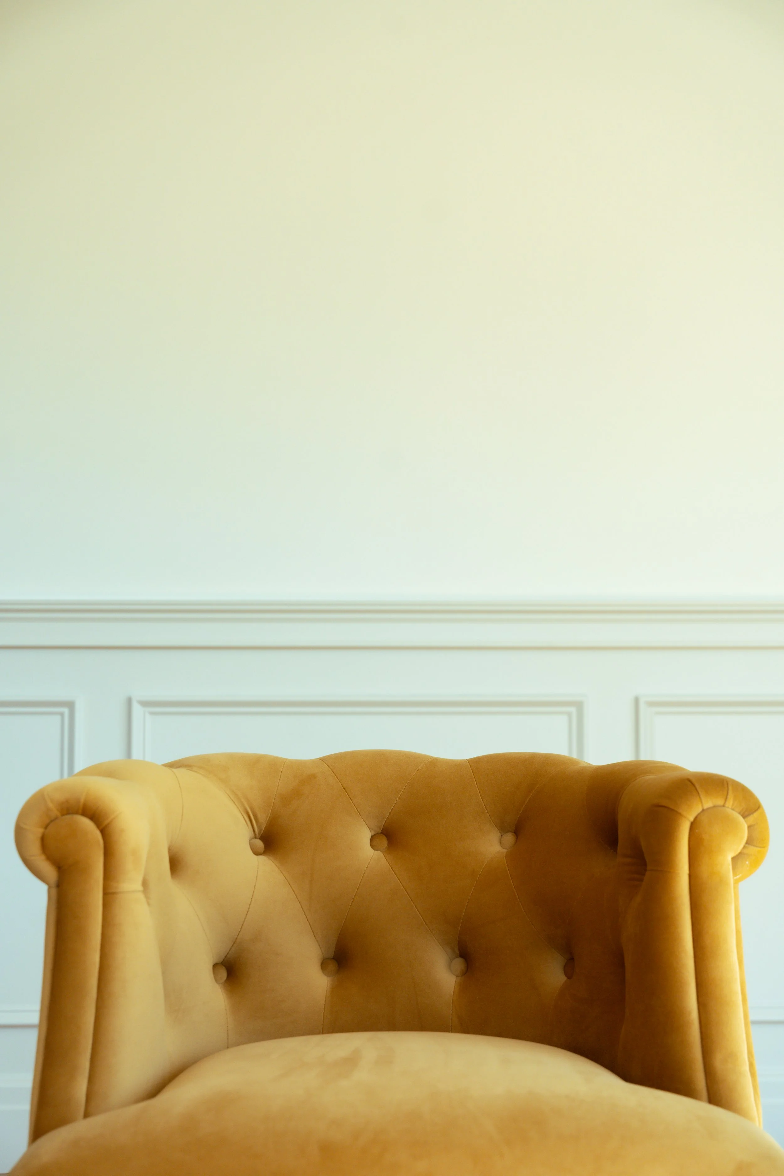 A yellow upholstered armchair with a tufted backrest in front of a white wall with decorative molding.
