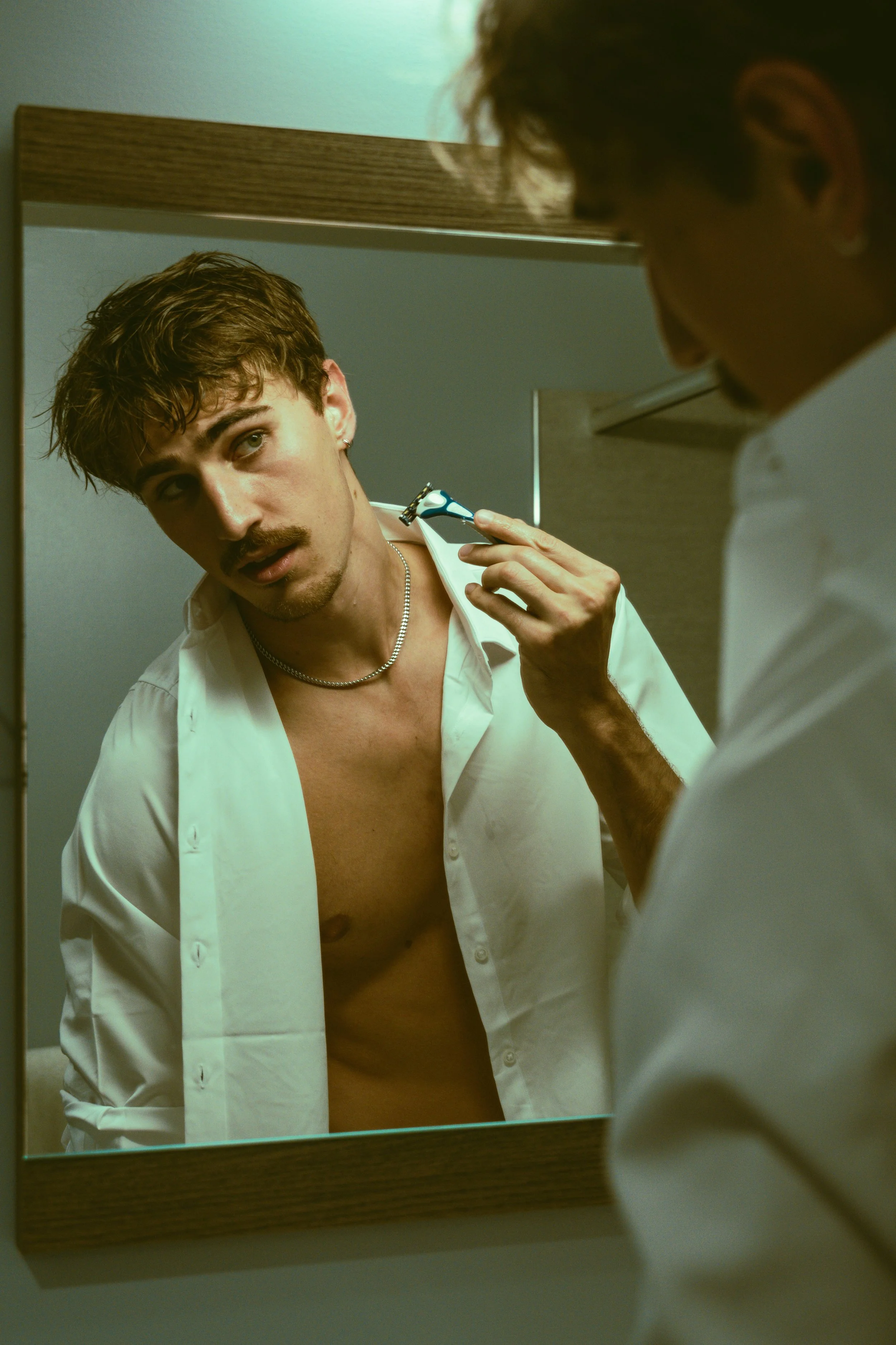A young man with curly hair and a mustache, topless and wearing an open white shirt, is looking into a mirror while trimming his facial hair with an electric shaver. The mirror has a wooden frame, and the background wall is plain.