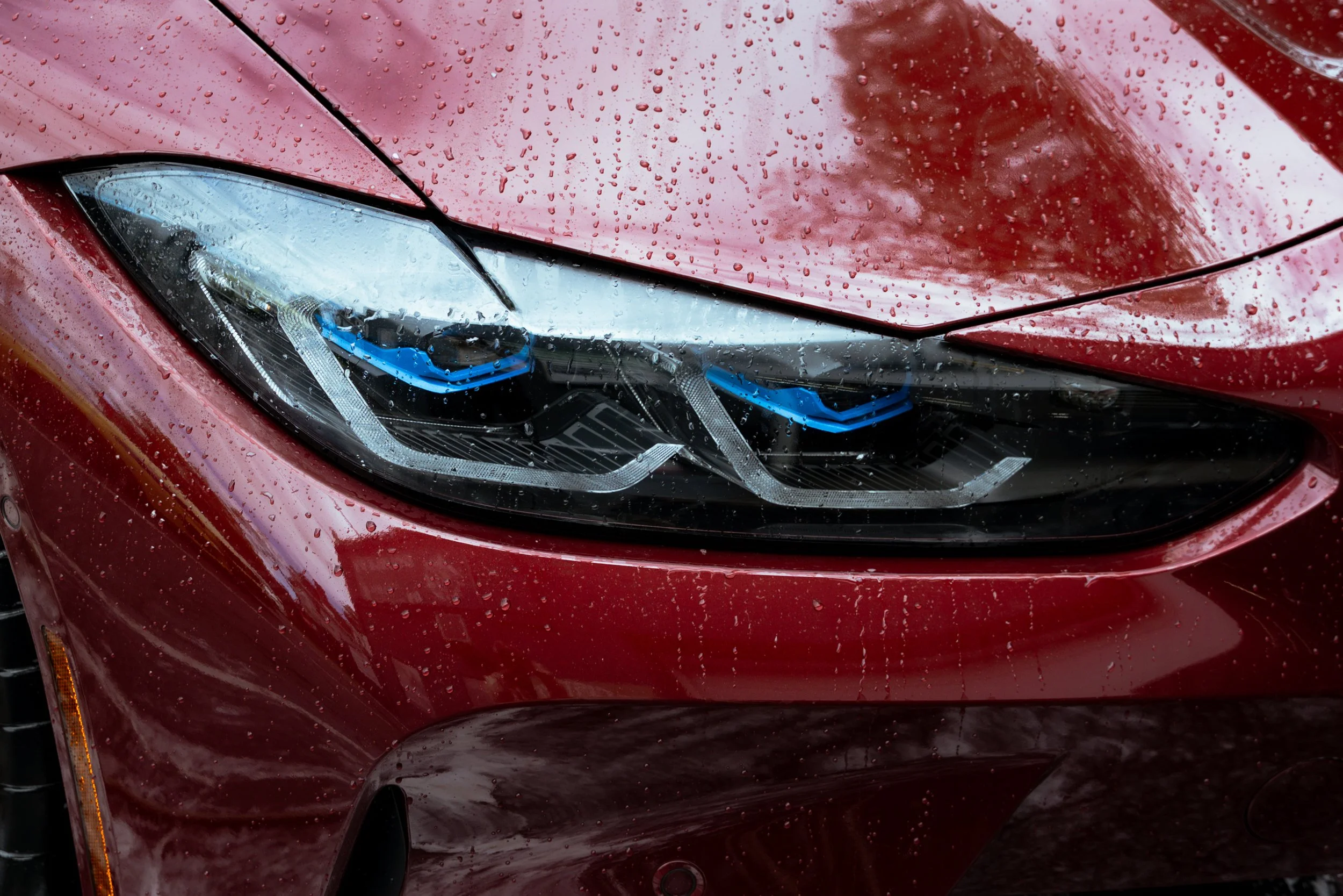 Close-up of a red car's front, showing the headlight and part of the hood with raindrops on the surface.