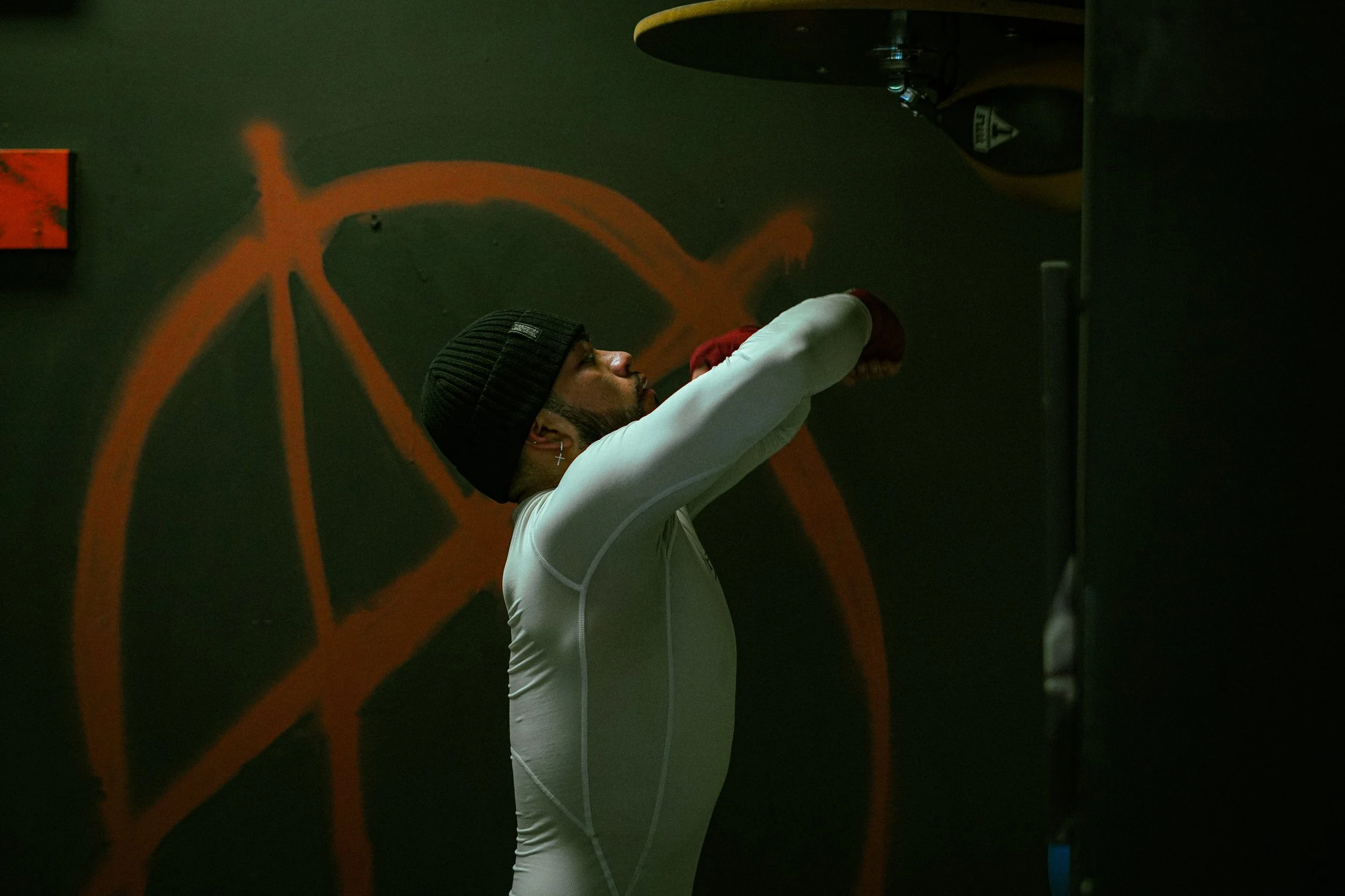 A man wearing a black beanie and white long-sleeve shirt doing cardio exercise in a gym, with an orange and black bike painted on the wall behind him.