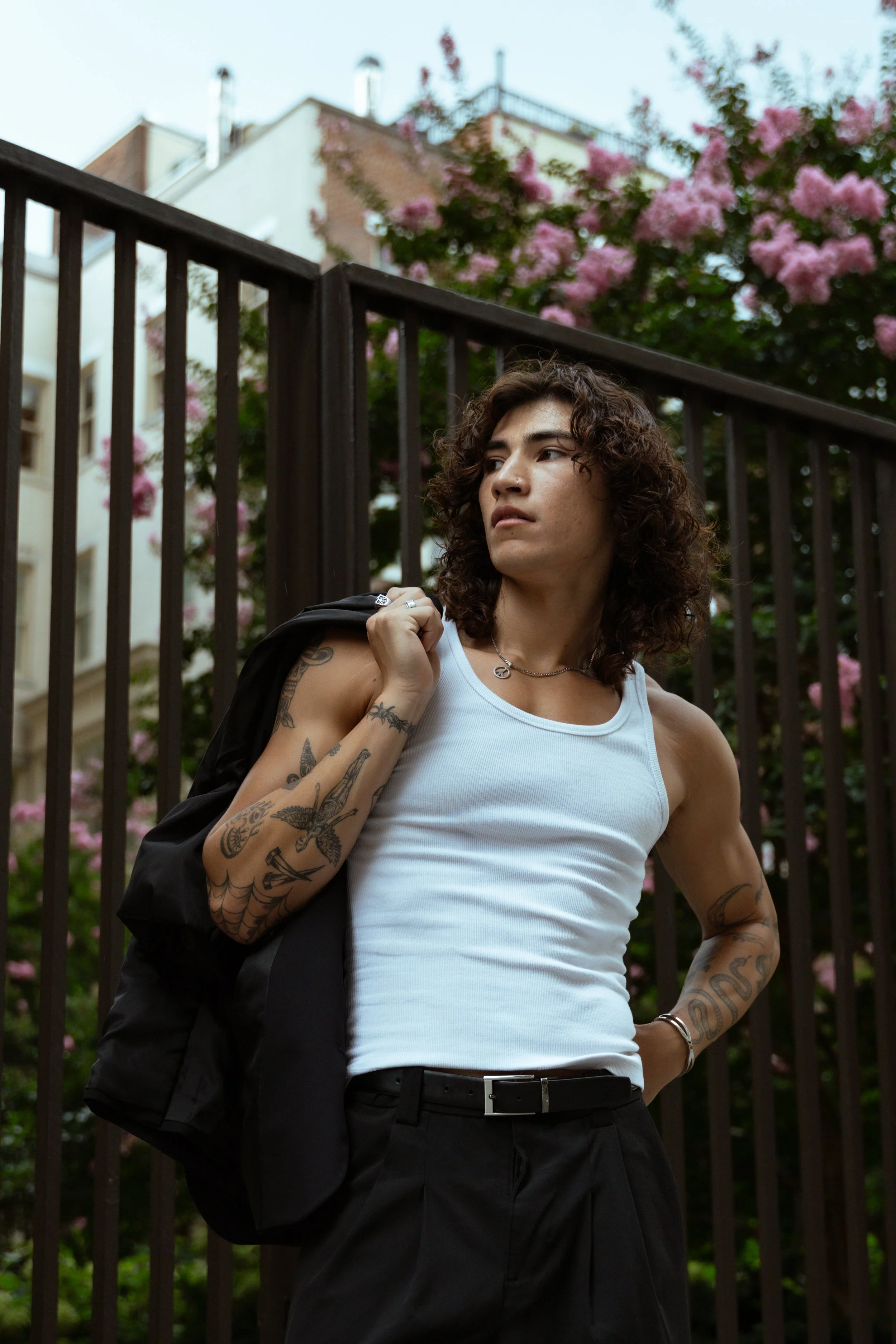 A person with curly brown hair, wearing a white tank top, black pants, and tattoos on their left arm and right forearm, stands outdoors near a black metal fence with pink flowering trees and buildings in the background, carrying a black jacket over t
