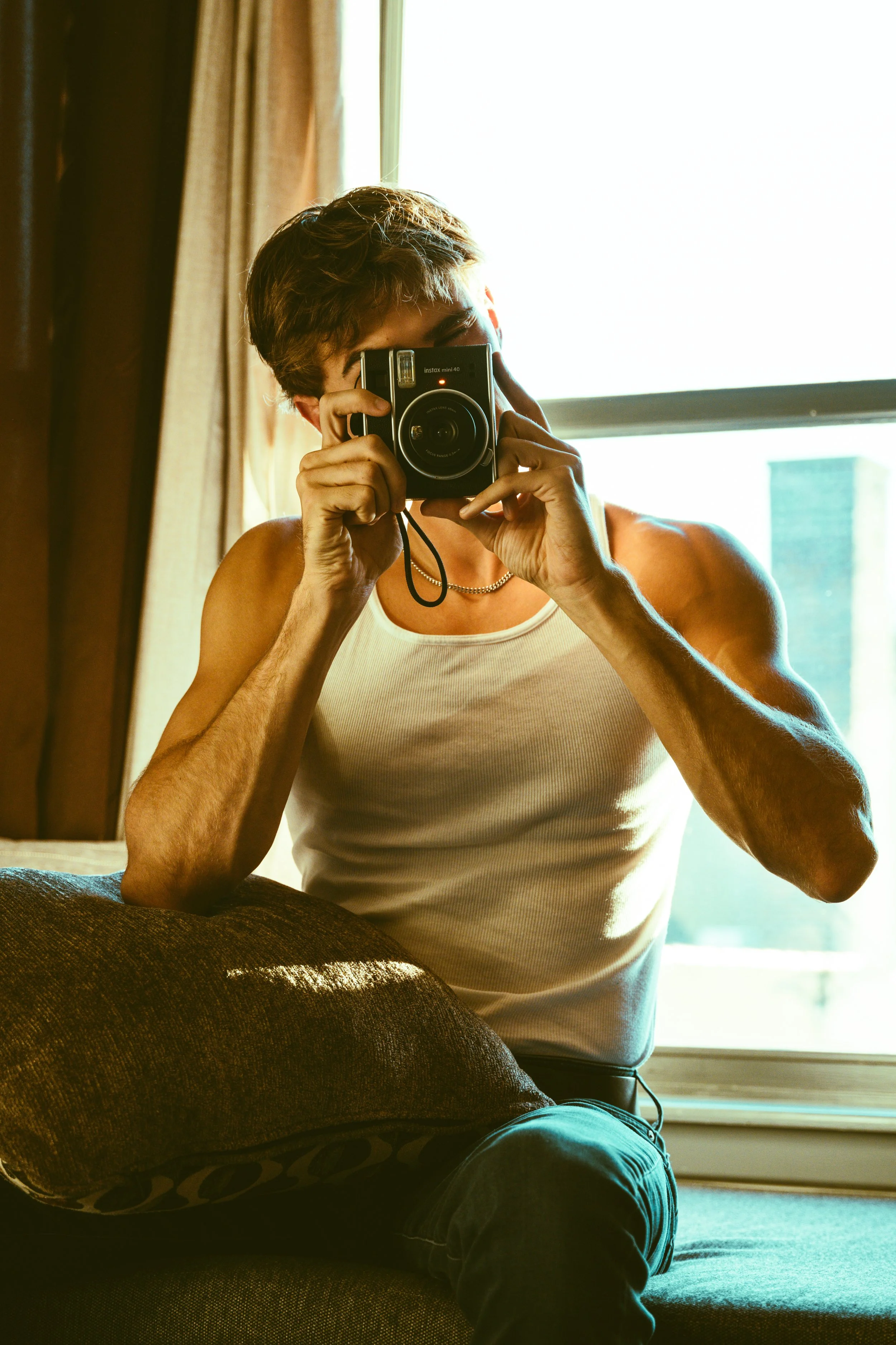 A person with short dark hair taking a mirror selfie with a vintage camera inside a room with natural light coming through a window.