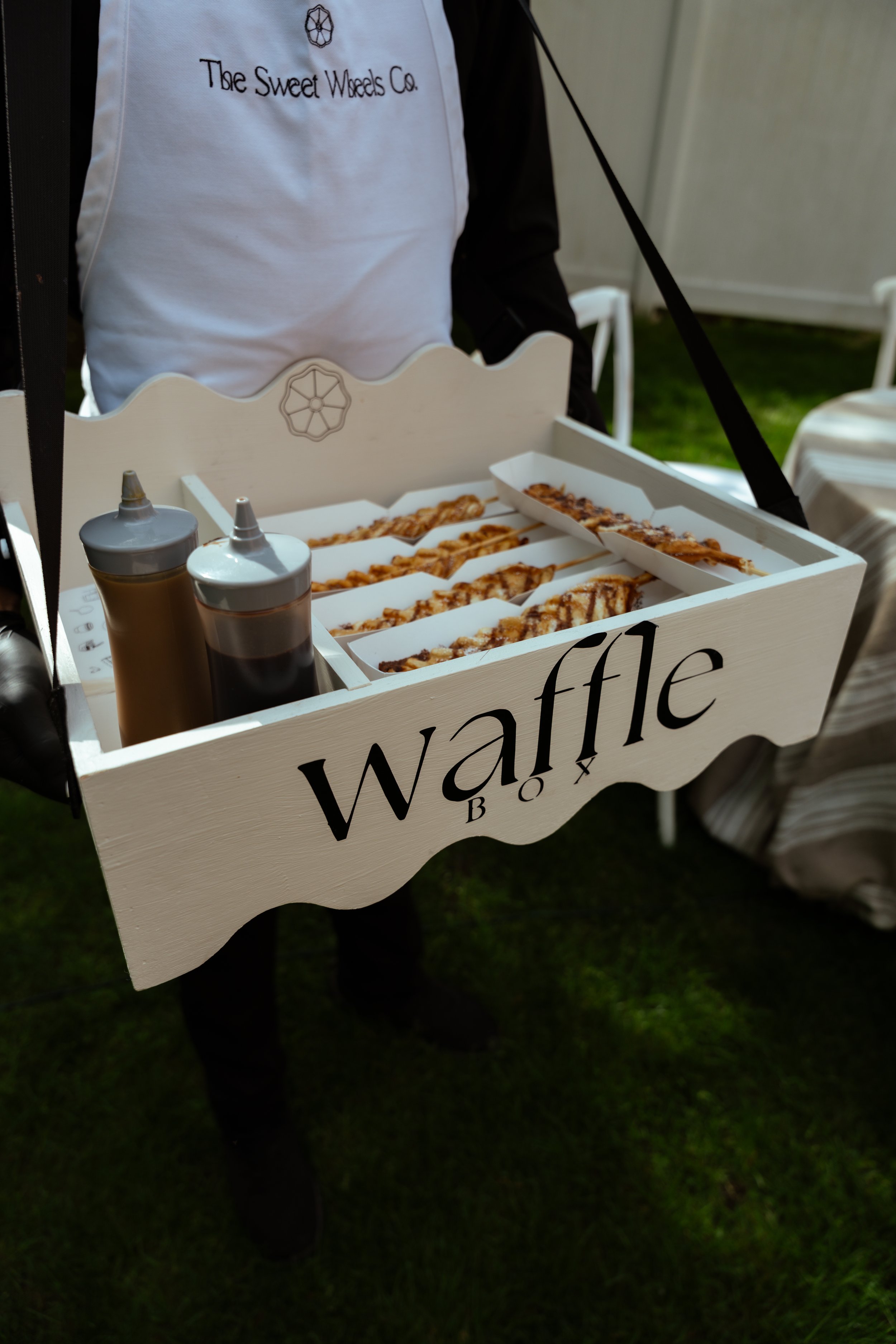 A person is holding a white wooden tray with the words 'waffle' written on it. The tray contains several waffle skewers drizzled with chocolate and caramel sauces. Two squeeze bottles with sauces are also on the tray. The person is wearing a white ap