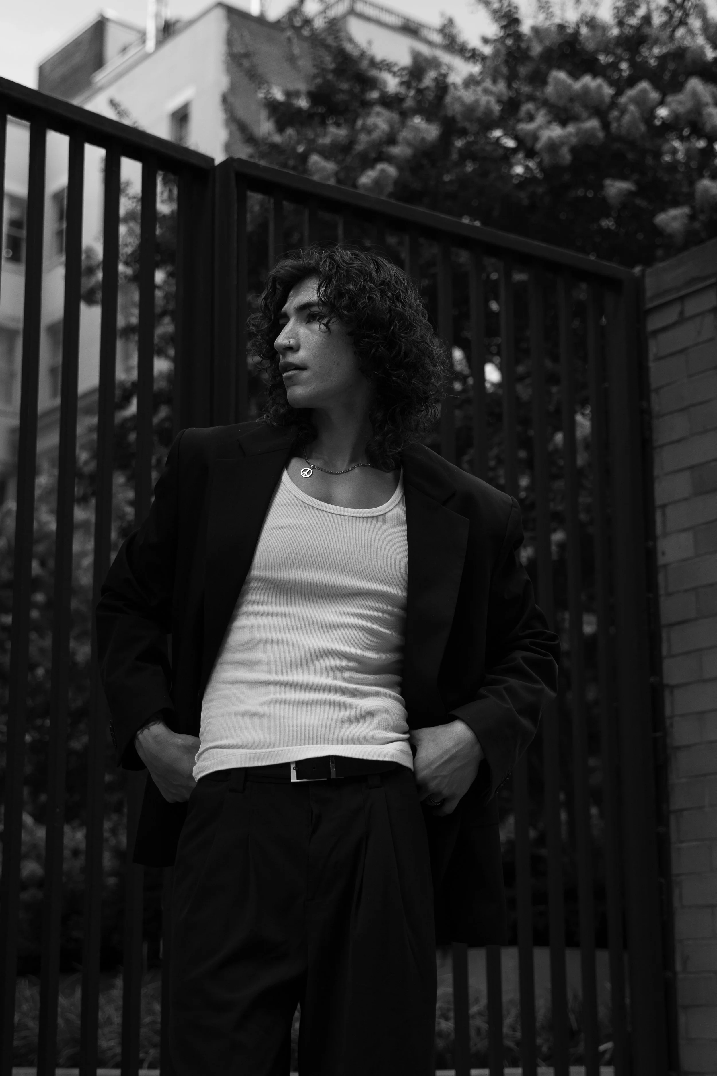 A woman with curly hair standing outdoors in front of a metal fence and a tree, wearing a blazer, a white tank top, and a peace symbol necklace.