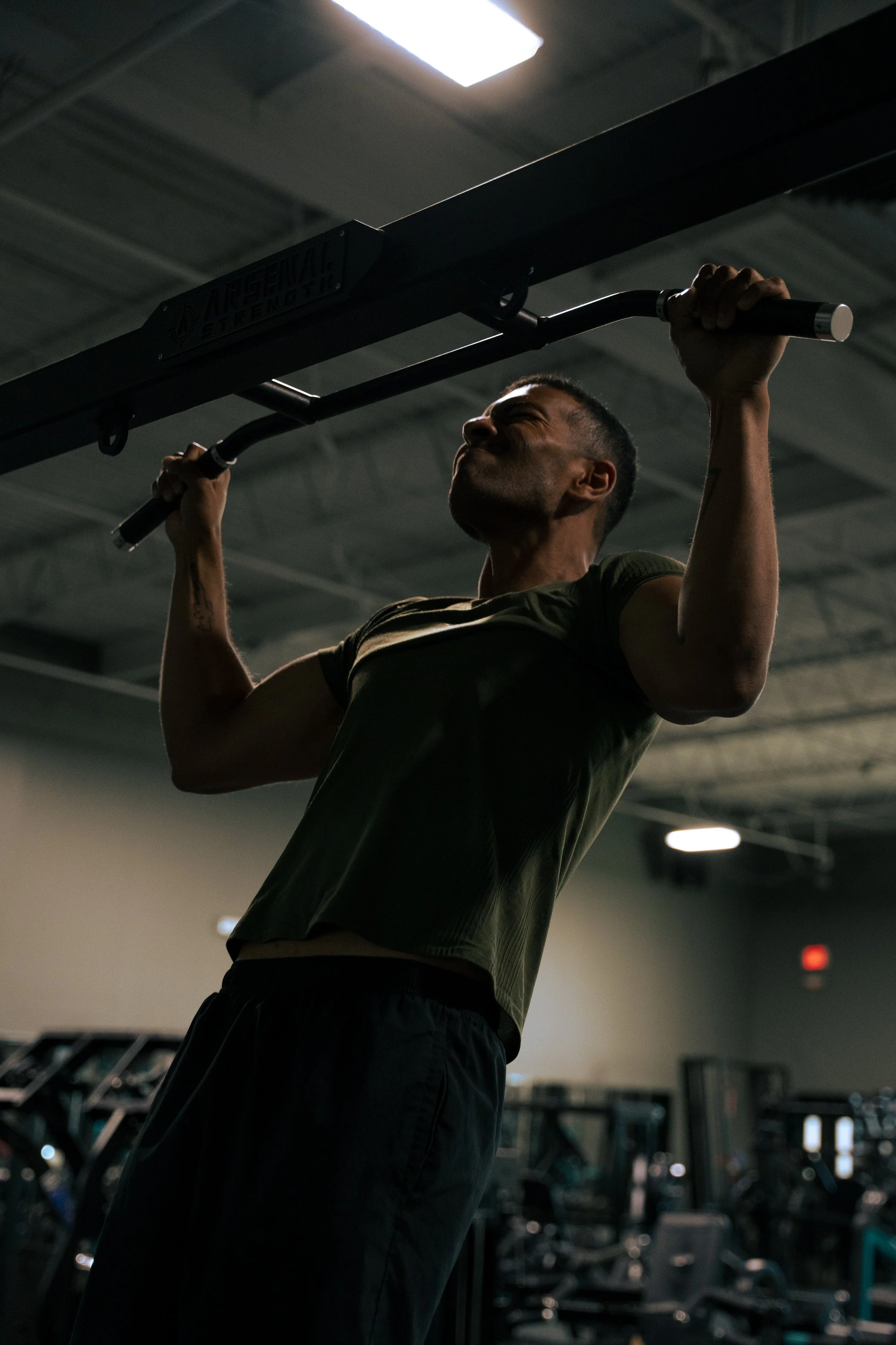 A man lifting a barbell in a gym, with focused expression, in a dimly lit setting.