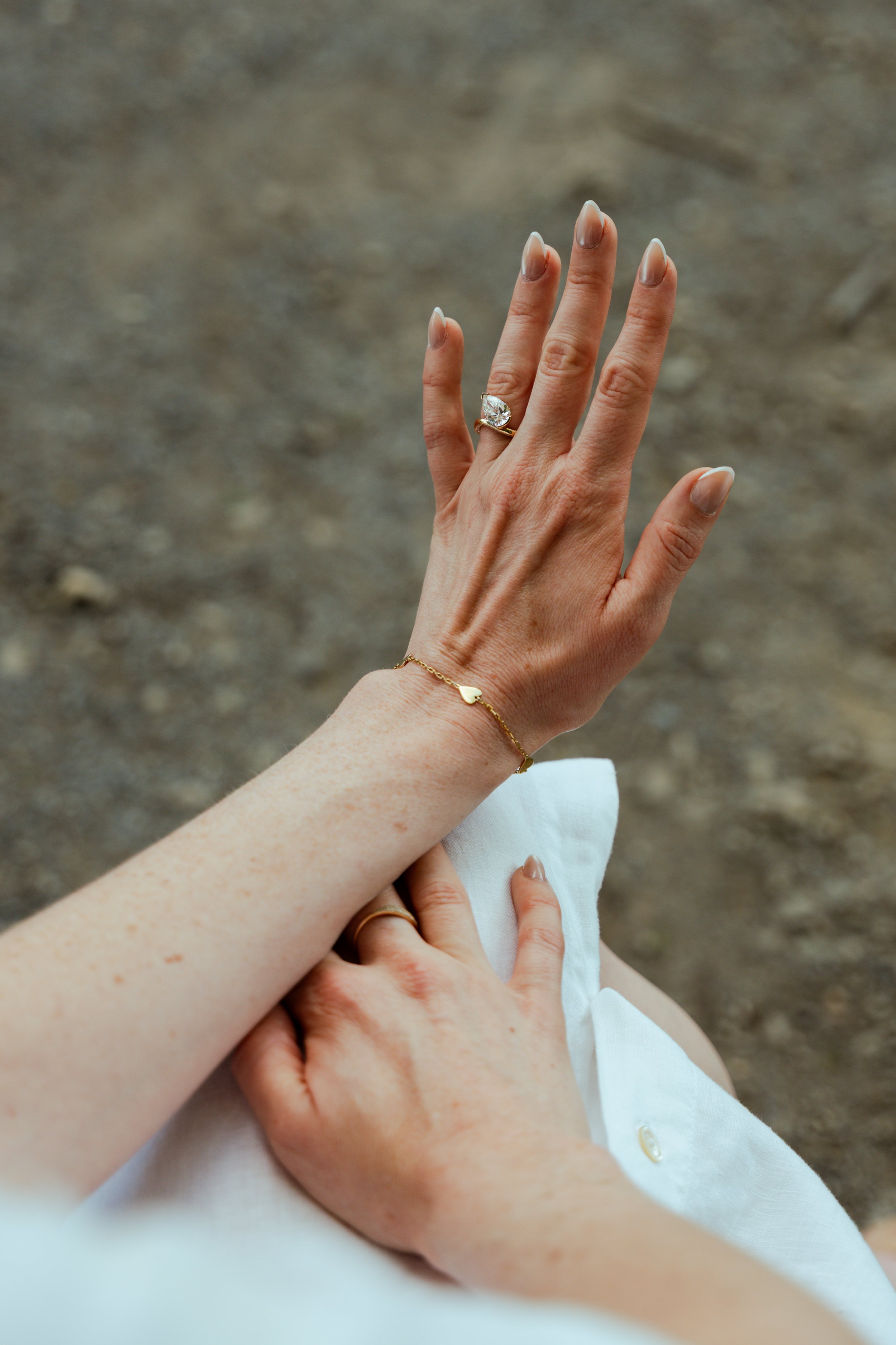 Close-up of a woman’s left hand showing a large, oval-shaped diamond engagement ring on her ring finger. The background is blurred and appears to be outdoors with a dirt ground. The woman is wearing a gold bracelet with a heart charm. Part of another