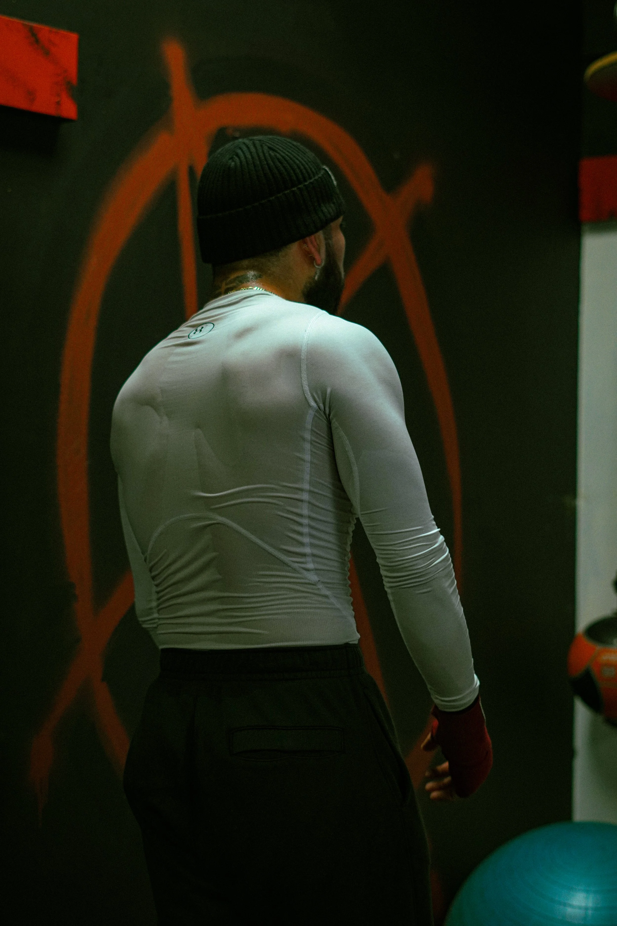 Rear view of a person wearing a black beanie, white long-sleeve athletic shirt, and black pants, standing in a gym with red and black painted walls and exercise equipment.
