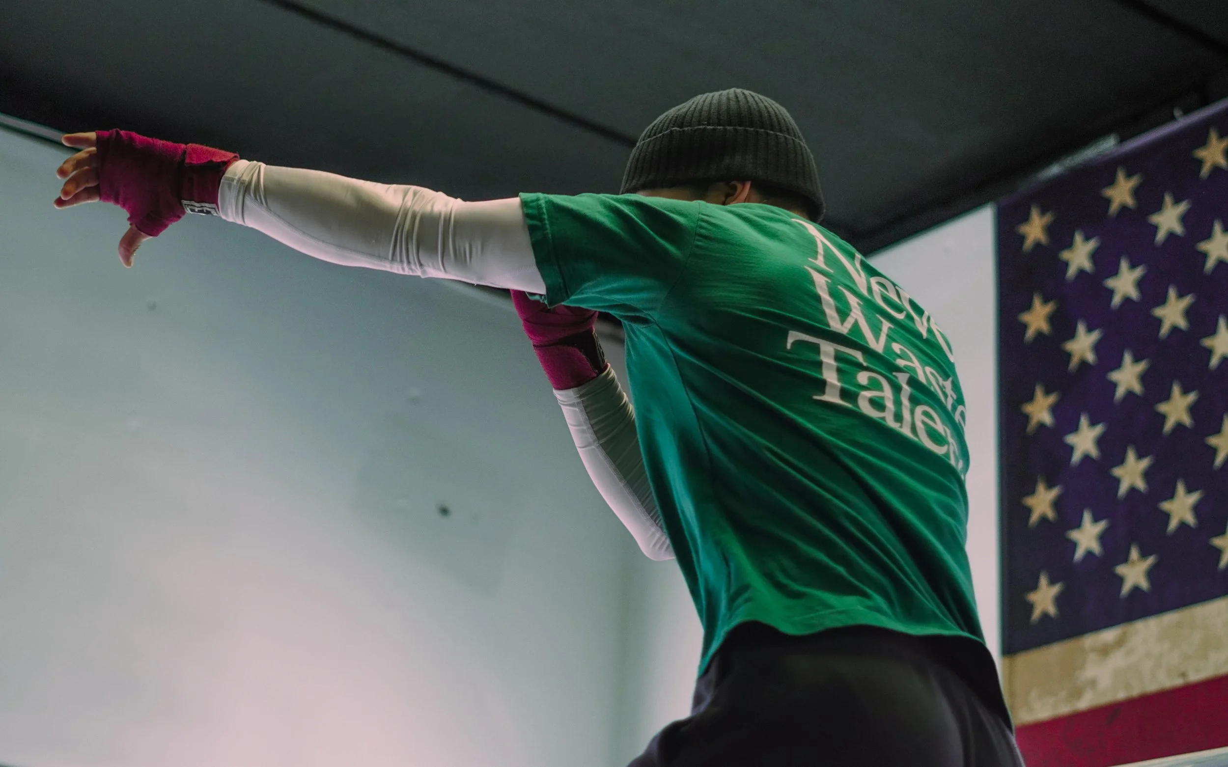 Person wearing a green t-shirt and a black beanie, stretching their right arm, in an indoor sports setting with an American flag in the background.