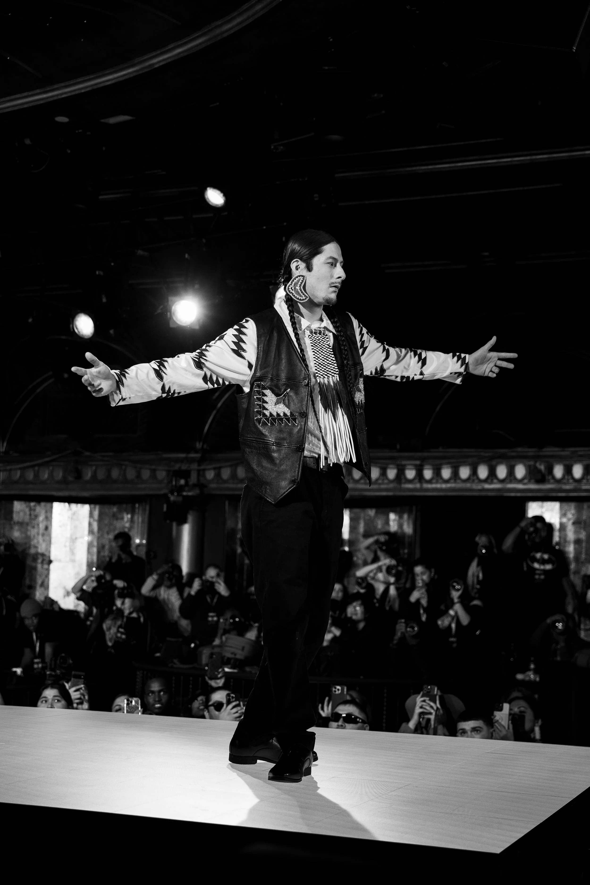 A performer with long braids, wearing a patterned shirt, leather vest, and large earrings, dancing on a stage with arms outstretched, in front of an audience taking photos.