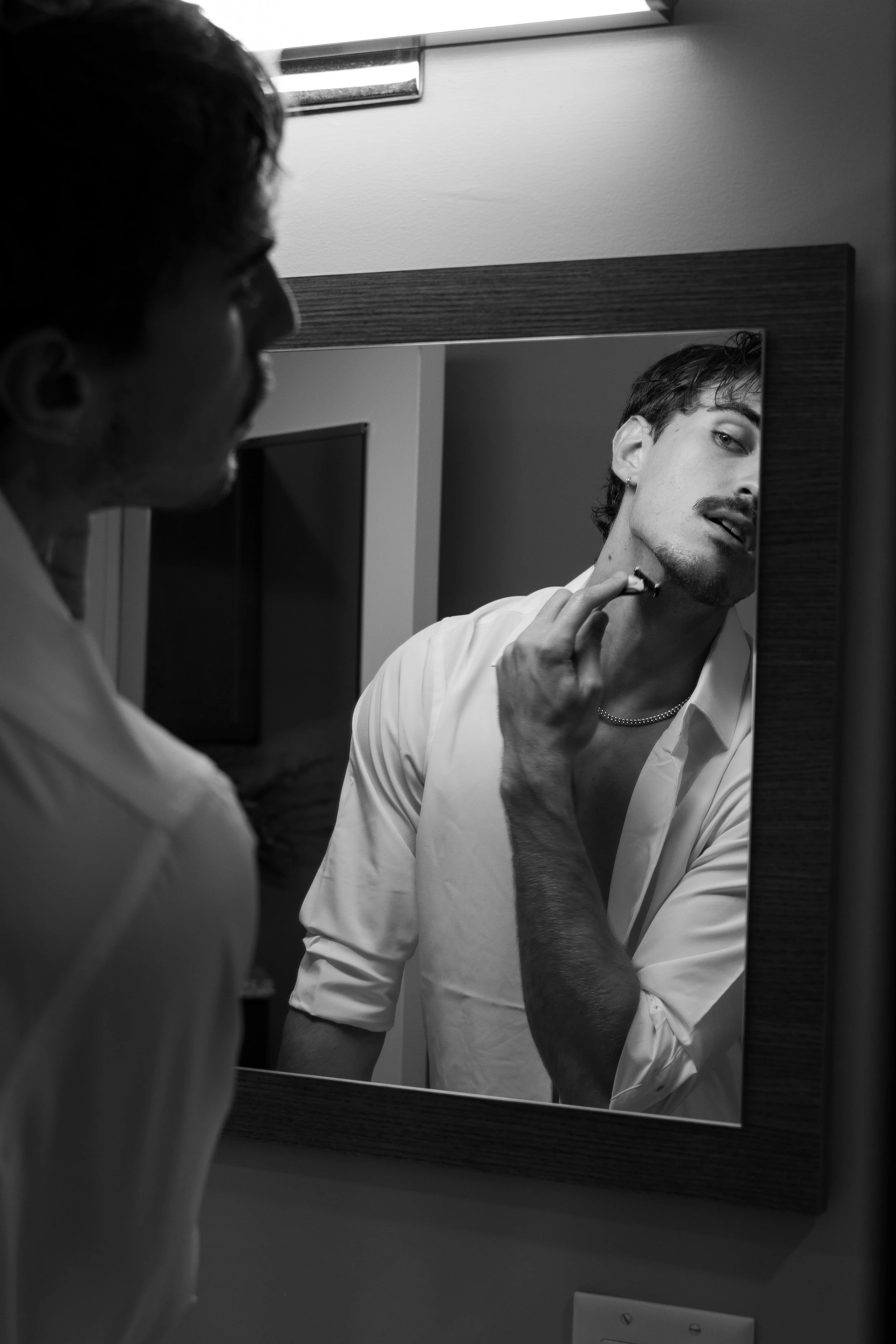 A young man with dark hair and a beard shaving in front of a mirror in a bathroom. He is wearing a light-colored shirt and a chain necklace, and he is holding a razor to his beard.