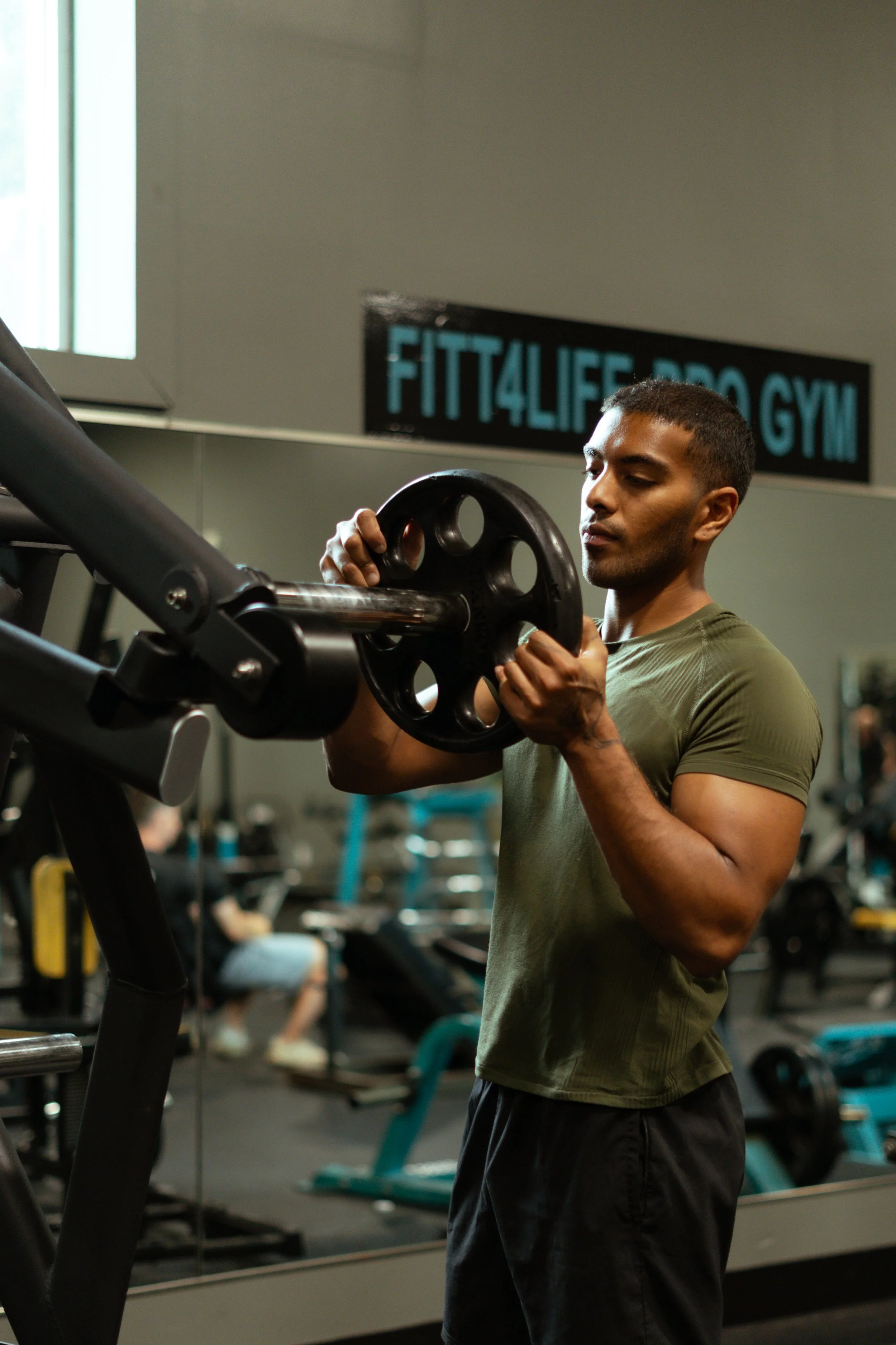 A man in a green t-shirt and black shorts holding a weight plate in a gym, with exercise equipment and a sign that reads 'FIT 4 LIFE PRO GYM' in the background.
