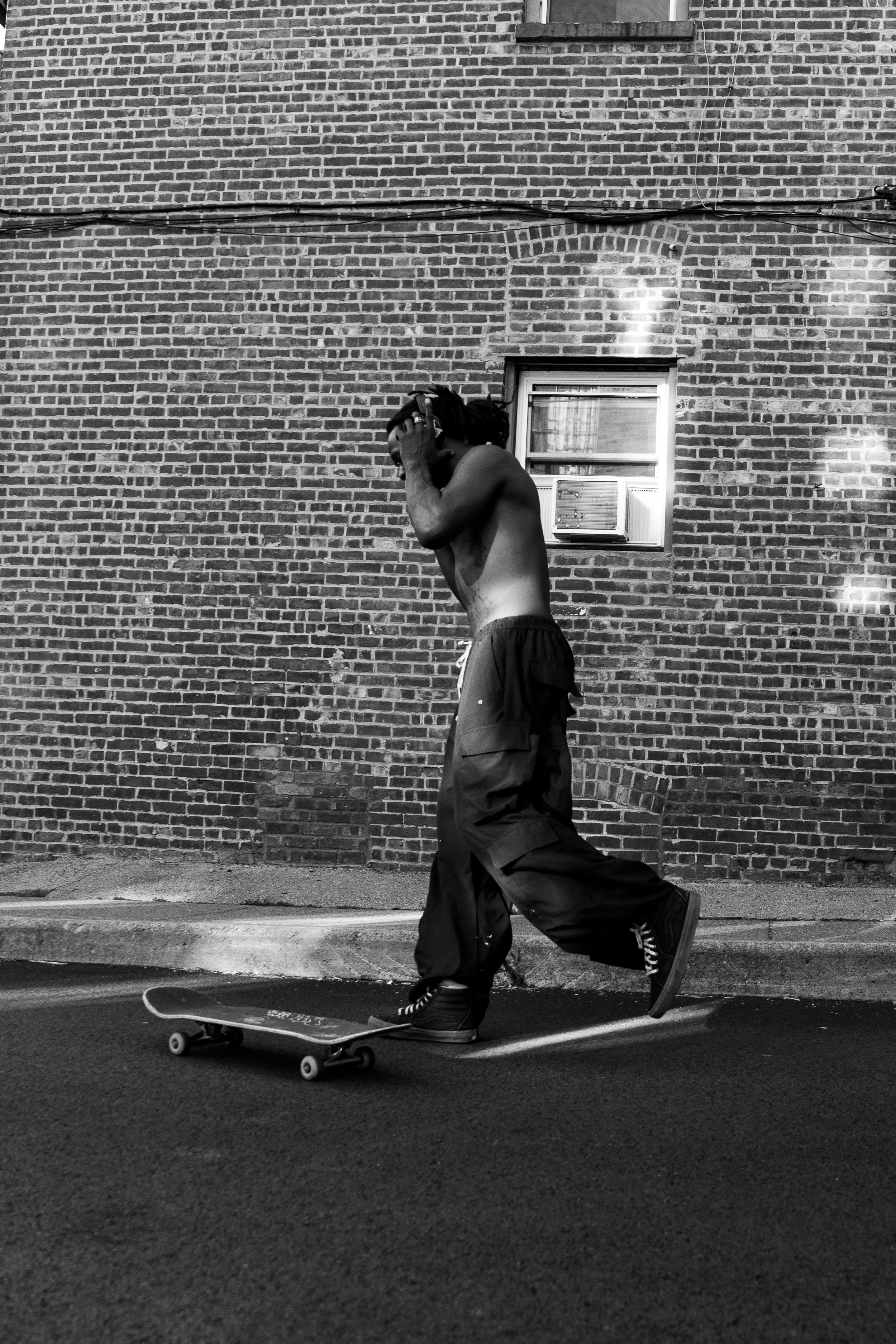 A shirtless young man with dreadlocks skateboarding along a street in front of a brick wall, with a window unit air conditioner visible in the wall.