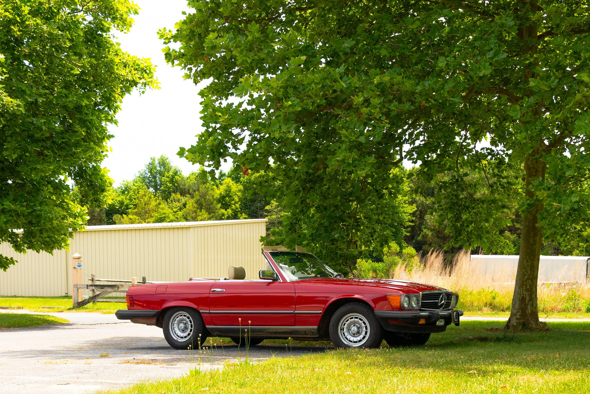 A red convertible car parked on a street with green trees and shrubs in the background under a partly cloudy sky.
