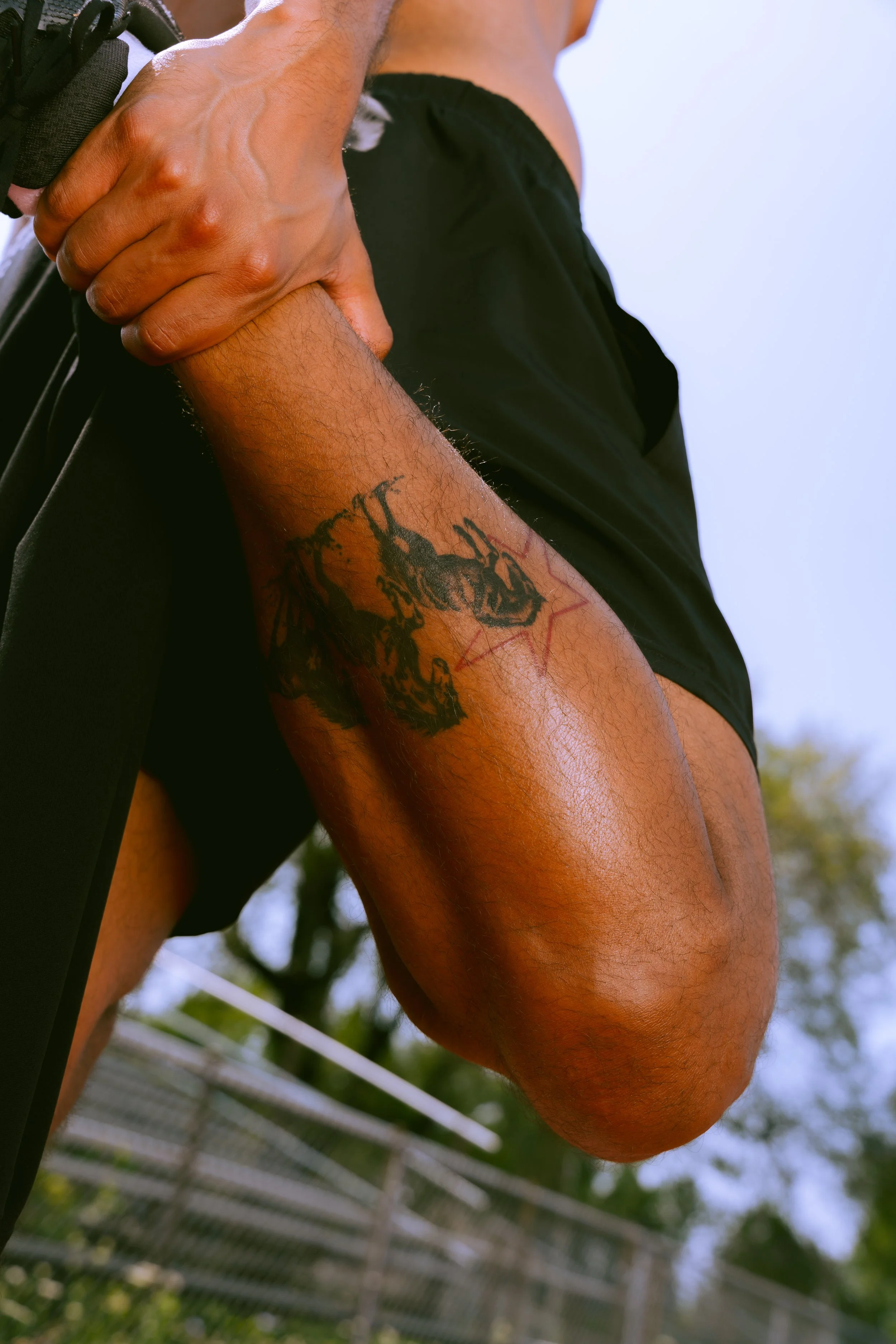 Close-up of a person with a tattoo on their forearm, holding their elbow with their hand, outdoors with a blurred background of trees and a fence.