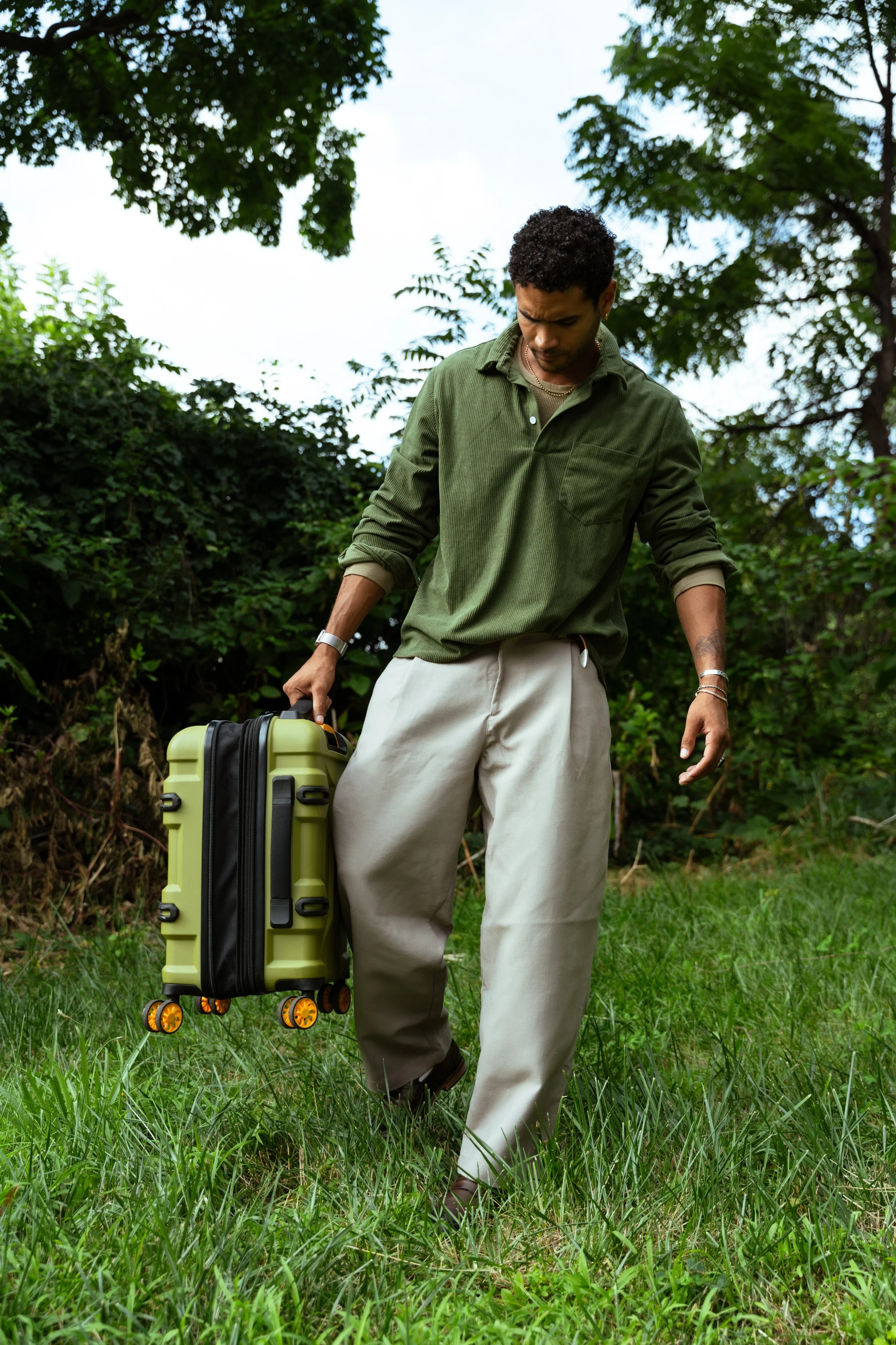 A person with curly hair wearing a green shirt and beige pants pulling a rolling suitcase on grass in a wooded outdoor area.