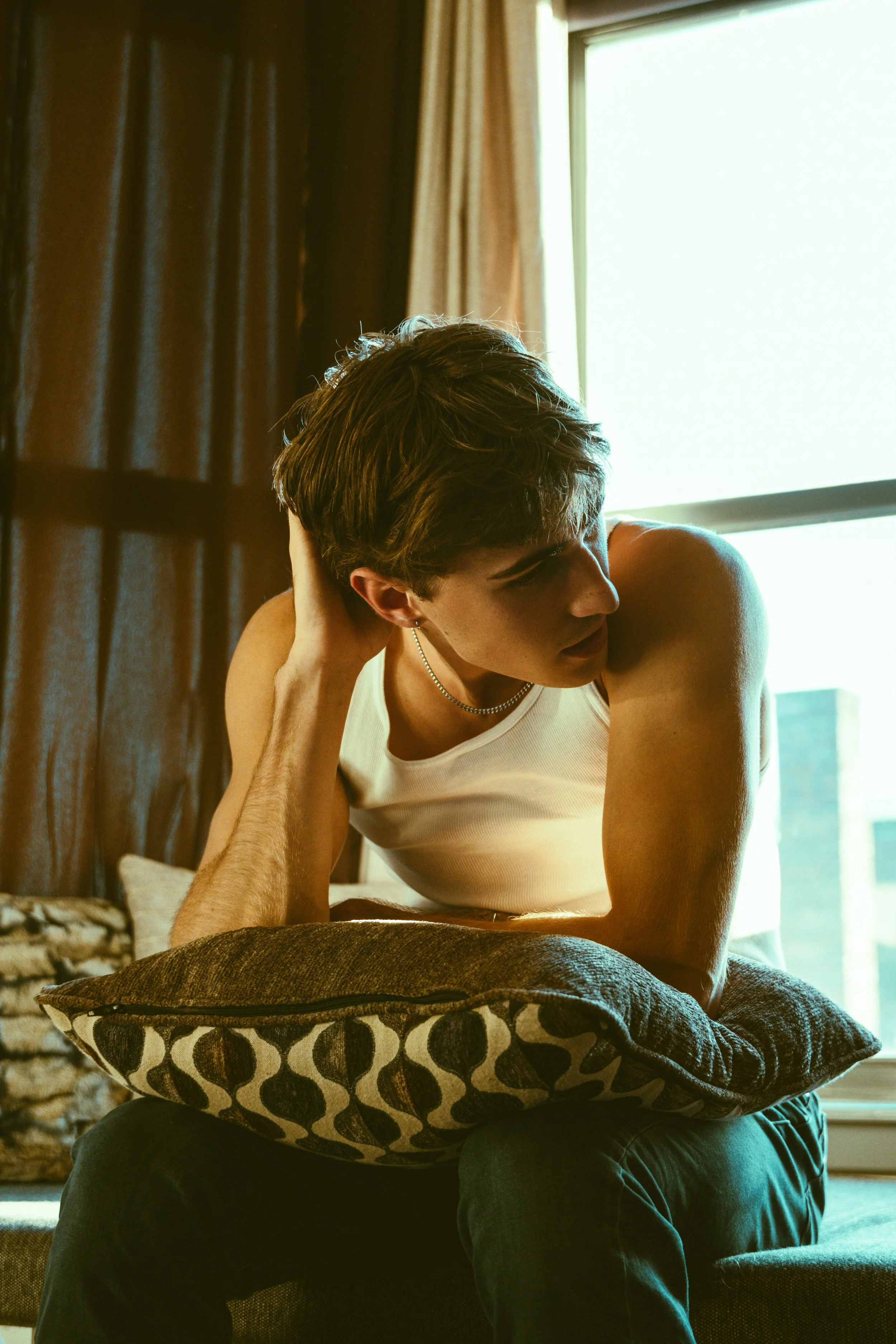 A young man with short brown hair wearing a white sleeveless shirt, sitting on a person's lap with a cushion supporting his arm, in front of a window with sunlight streaming in, and brown curtains in the background.
