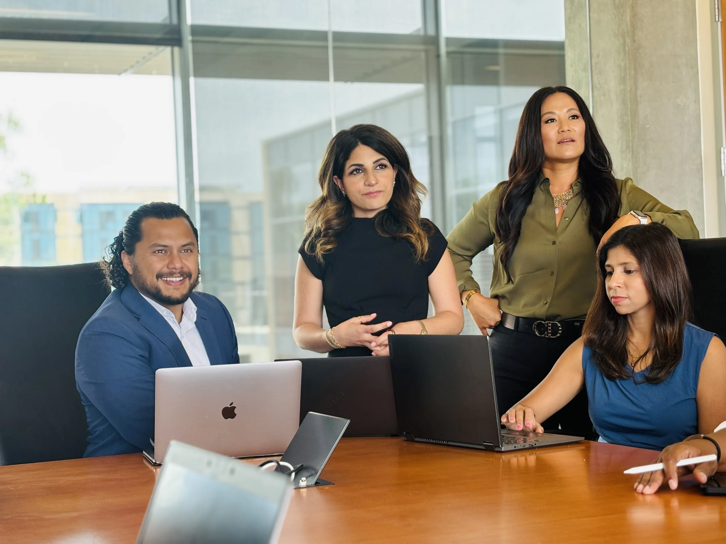 Four diverse business professionals engaged in a discussion in a modern office conference room, with laptops on the table.