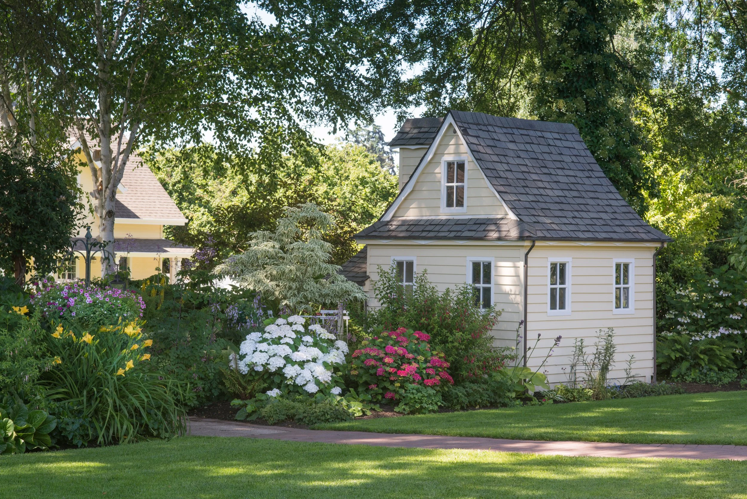 A well-maintained garden with vibrant flowers, lush green grass, and a cream-colored house with a dark gray roof, surrounded by trees.