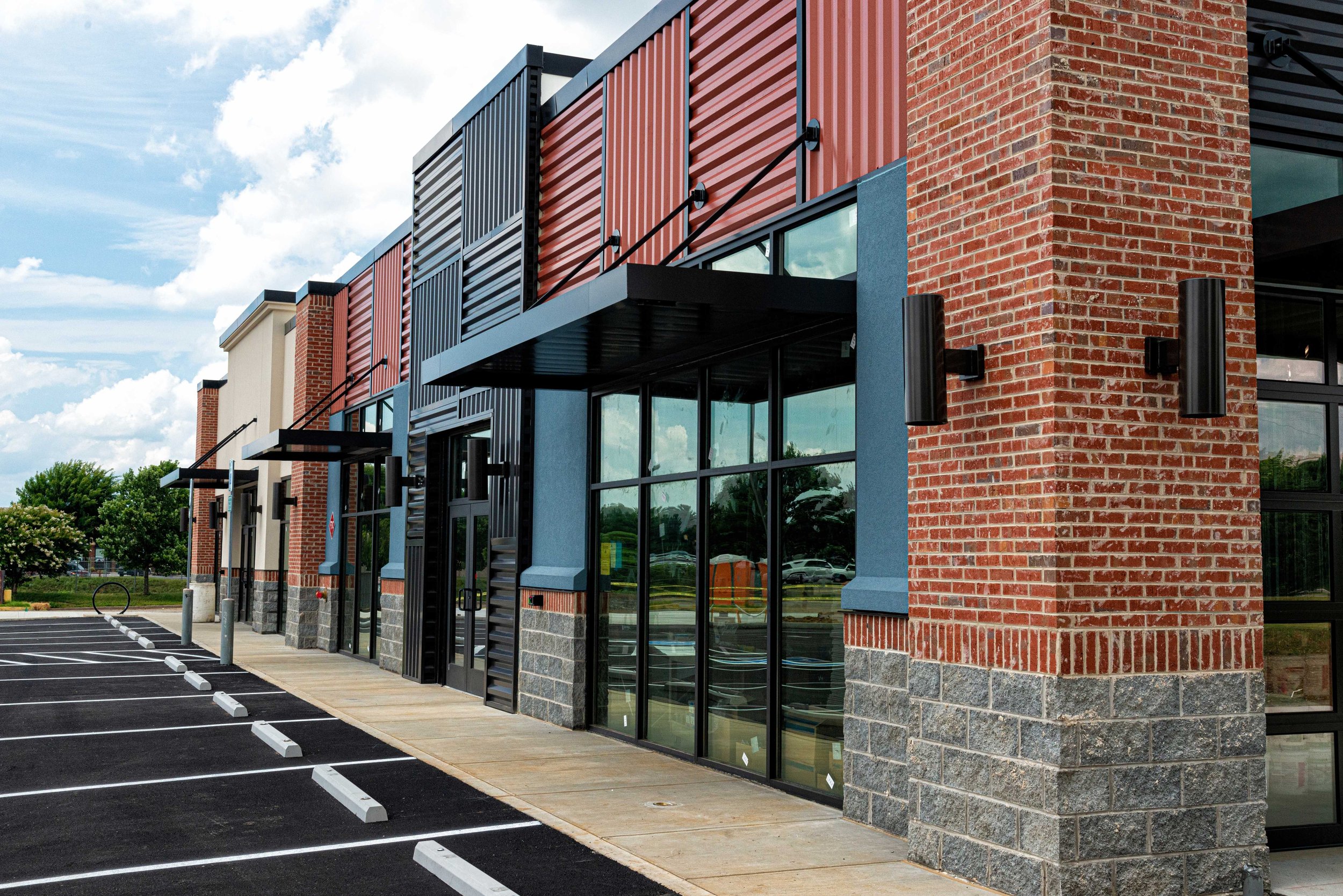 Modern commercial building with brick and metal exterior, large glass windows, and a small parking lot in front.