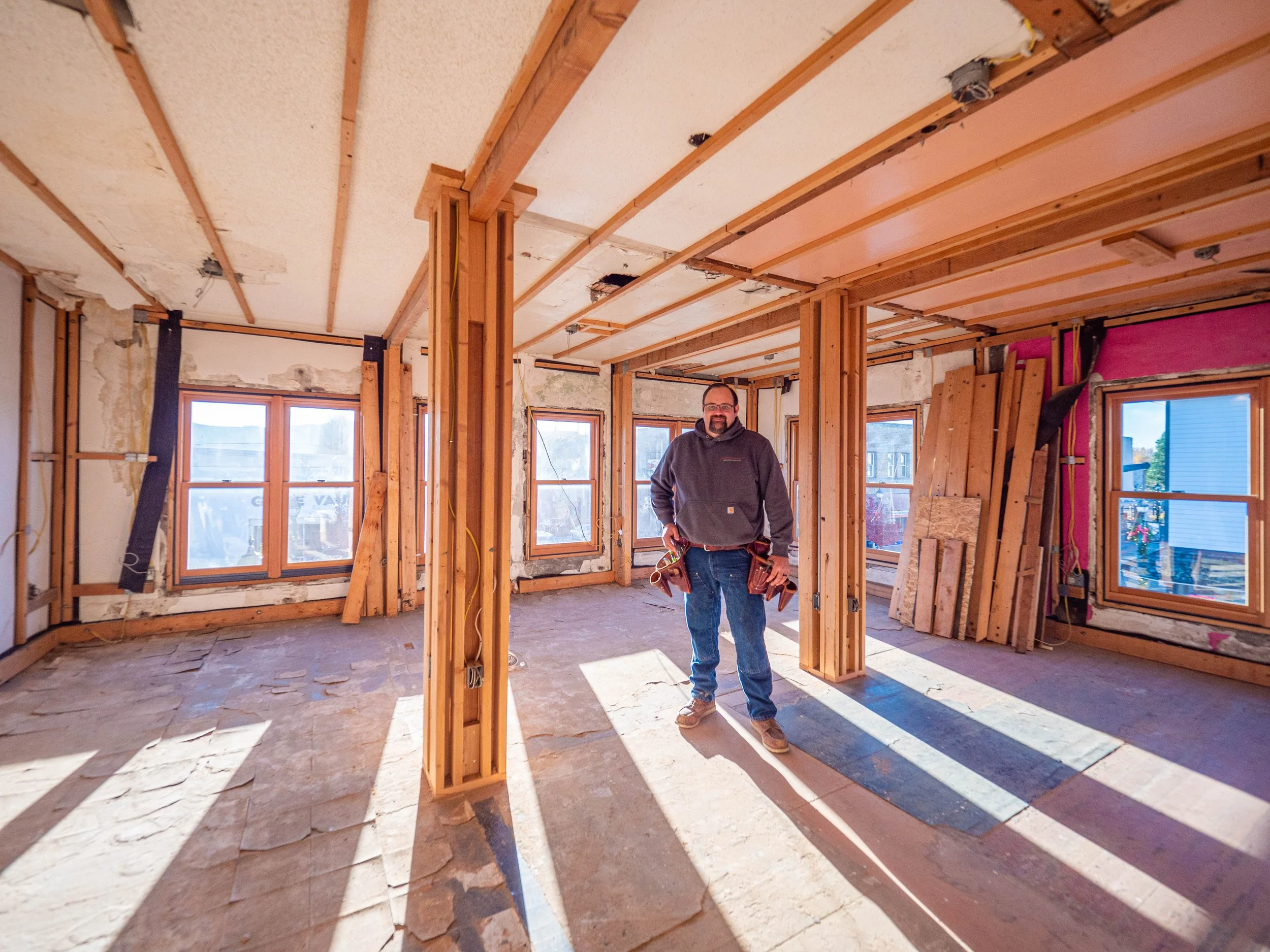A man standing inside a house under renovation, with exposed wooden framing, large windows, and construction materials on the floor.