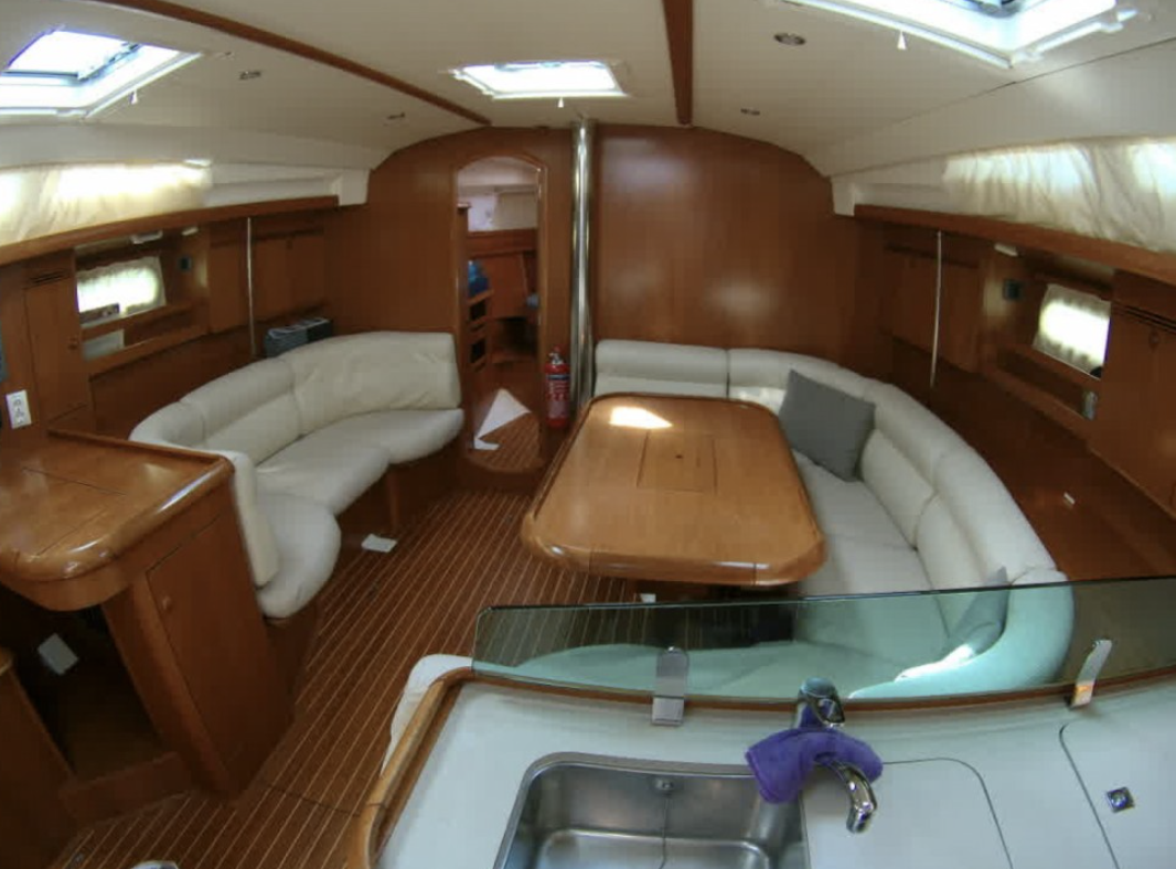 Interior of a yacht with wooden paneling, white cushioned seating, a dining table, and a kitchen area with a sink.