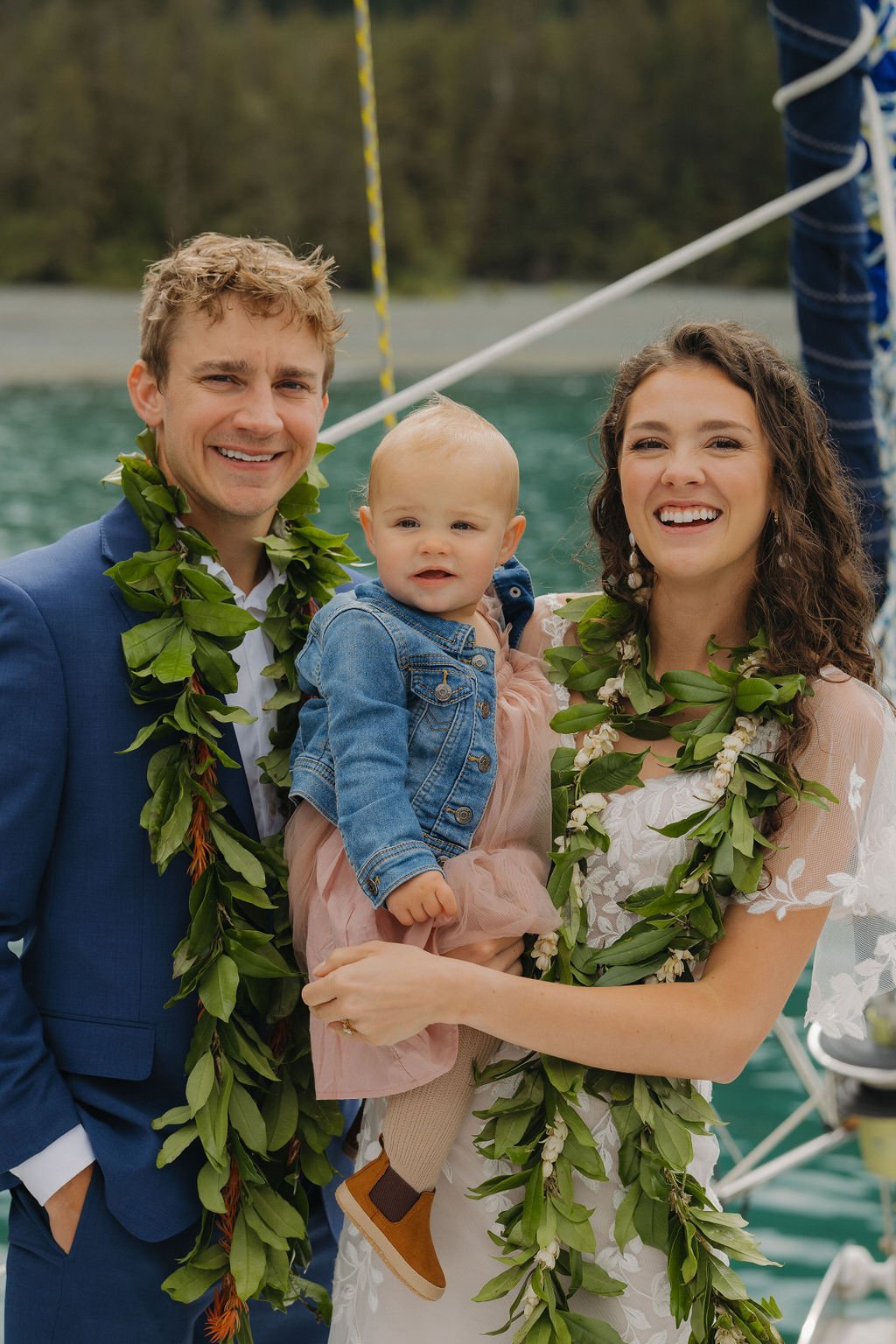 Happy couple with baby on a boat, wearing floral garlands and formal attire.