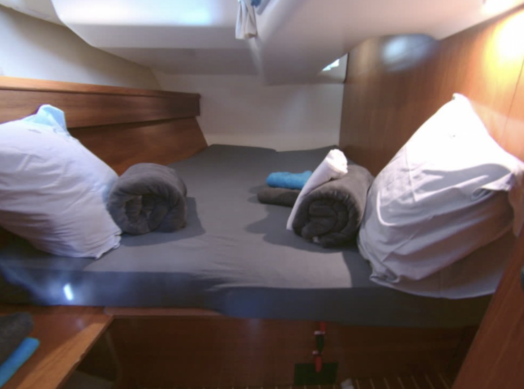 Interior of a small bedroom with a bed, pillows, and rolled towels, featuring wooden paneling and natural light.