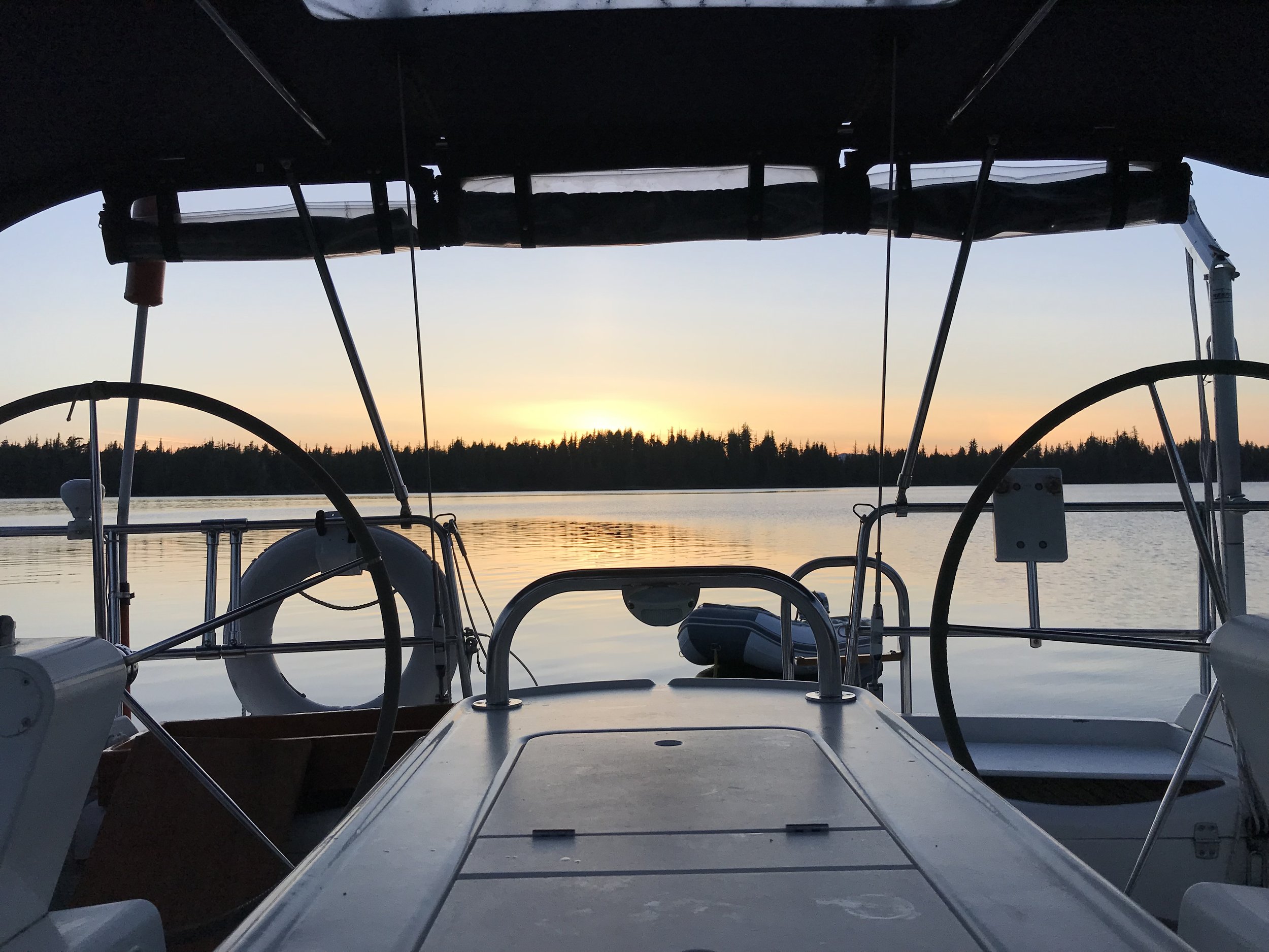 View from a yacht cockpit at sunset, with silhouette of steering wheels, life ring, and calm water reflecting the sky.
