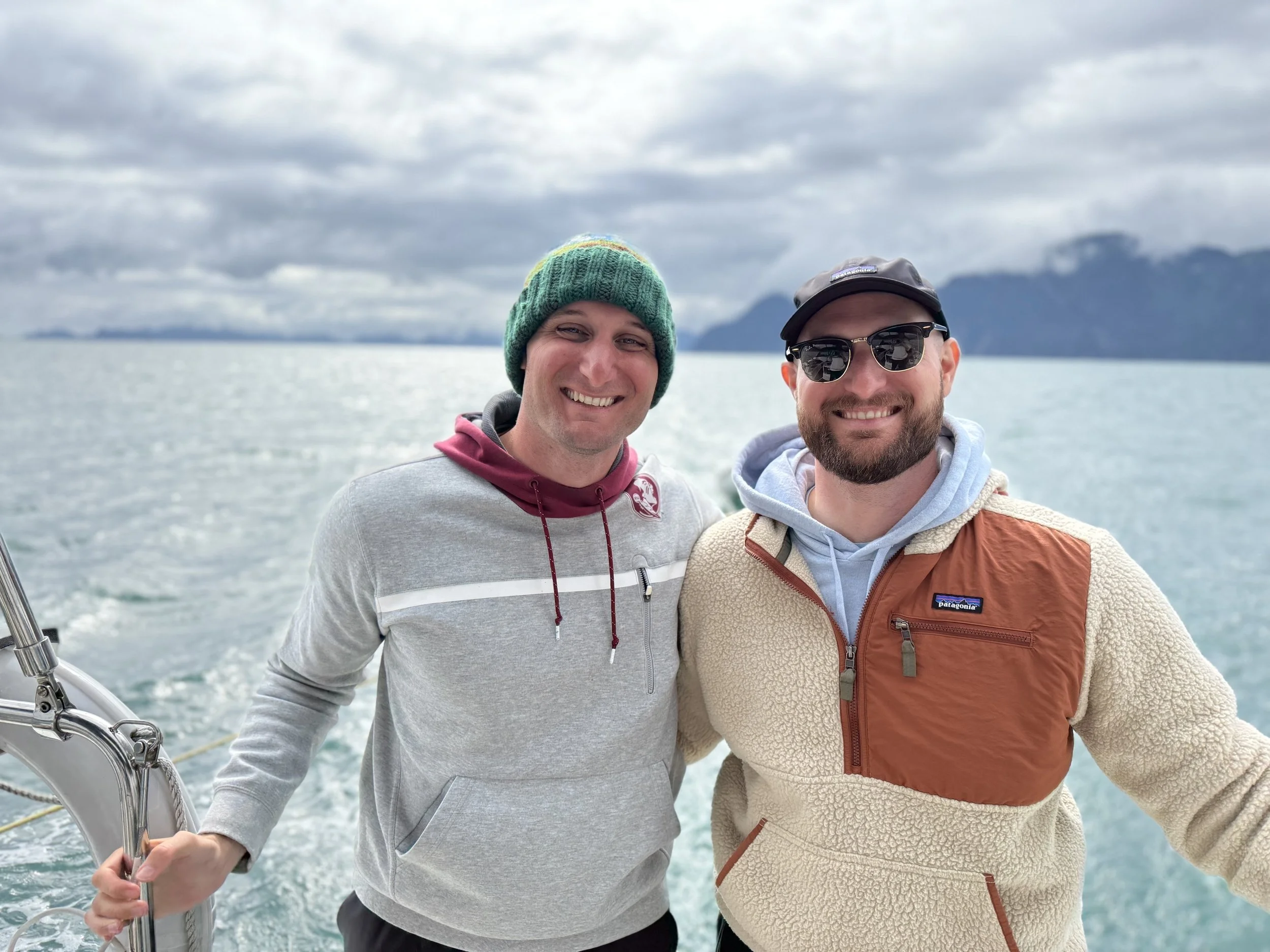 Two men smiling on a boat with water and mountains in the background, dressed in casual outdoor clothing.