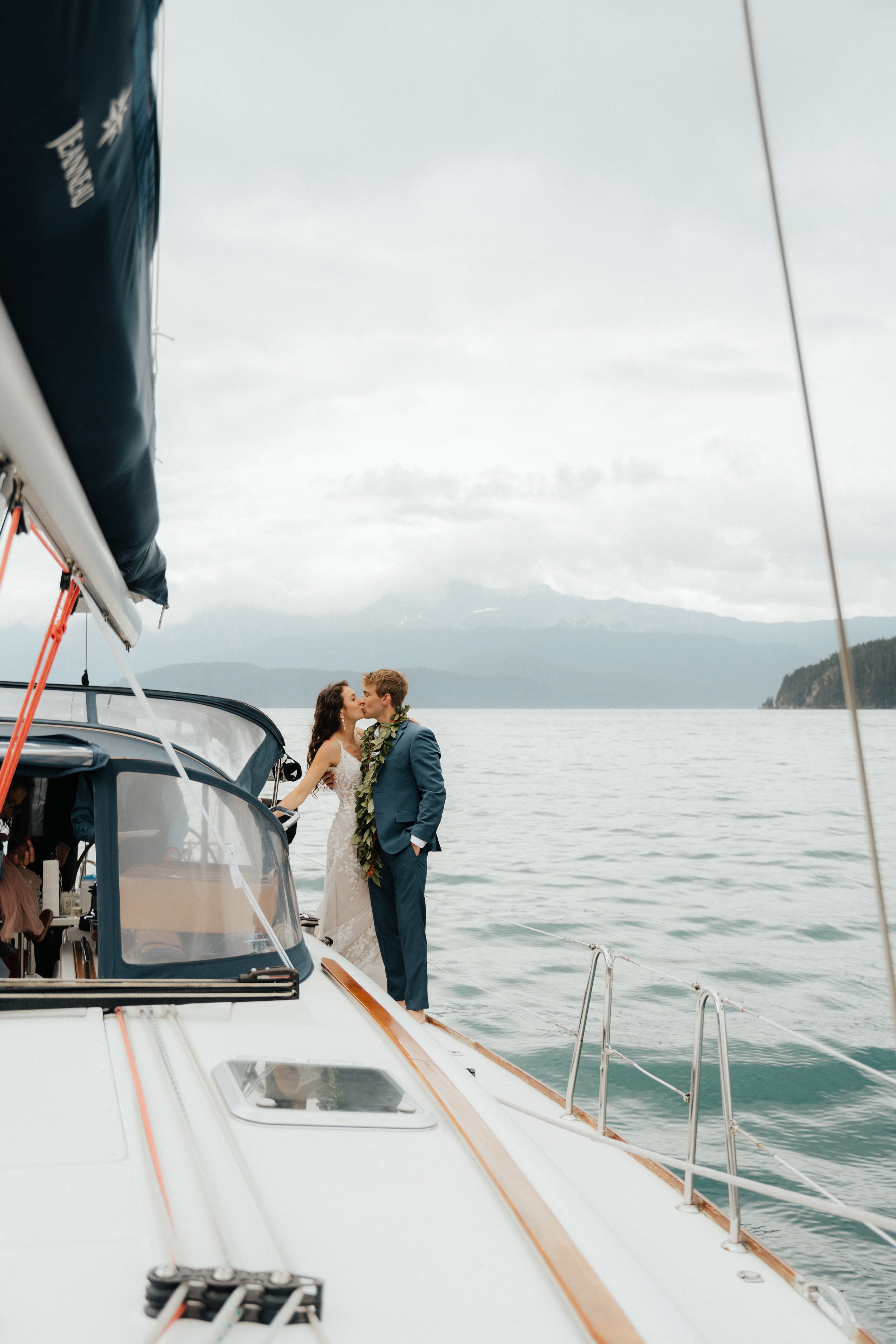 A couple kissing on a sailboat with a scenic mountain and water background.