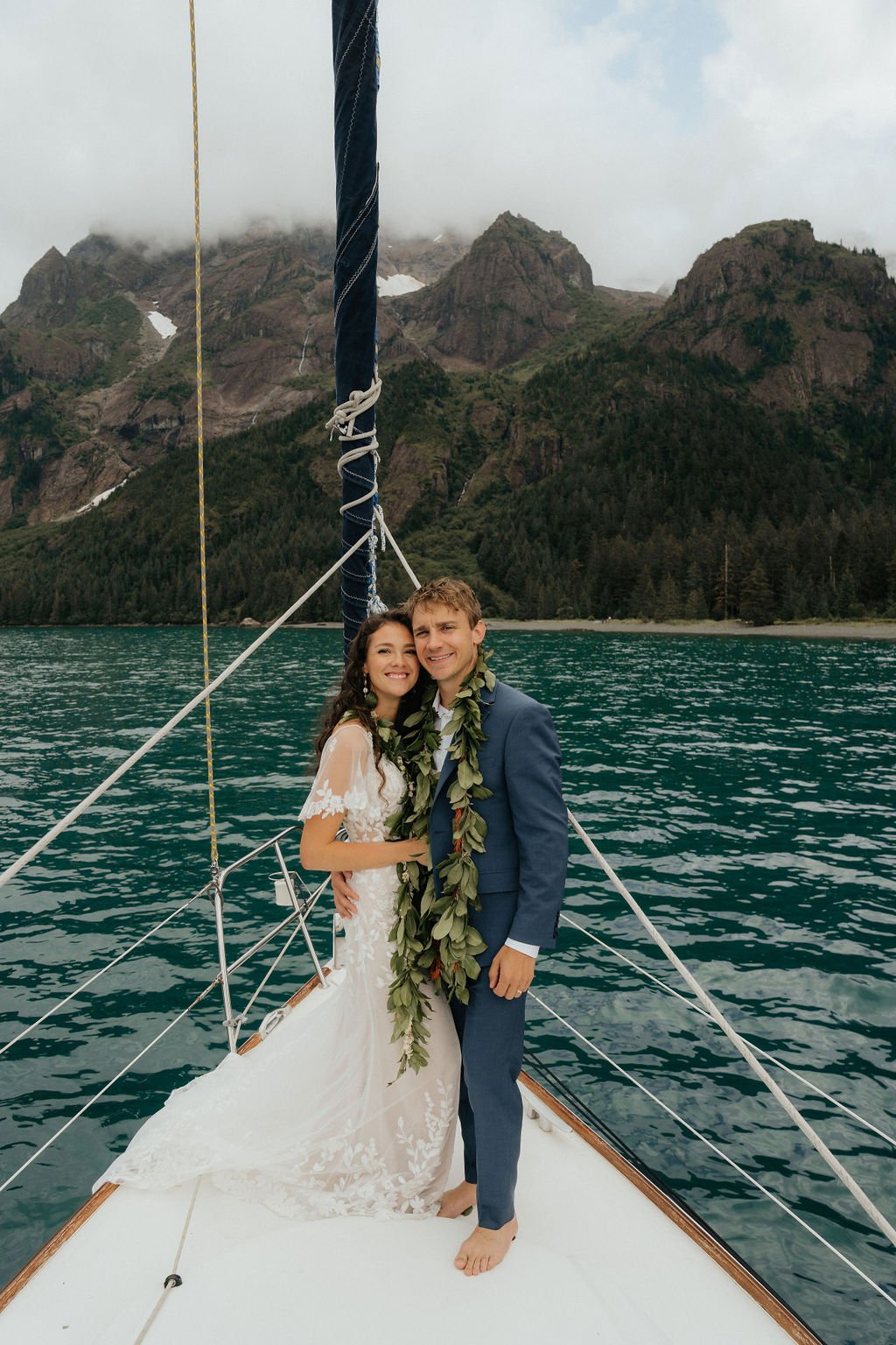 Couple in wedding attire on a sailboat with mountains in the background.