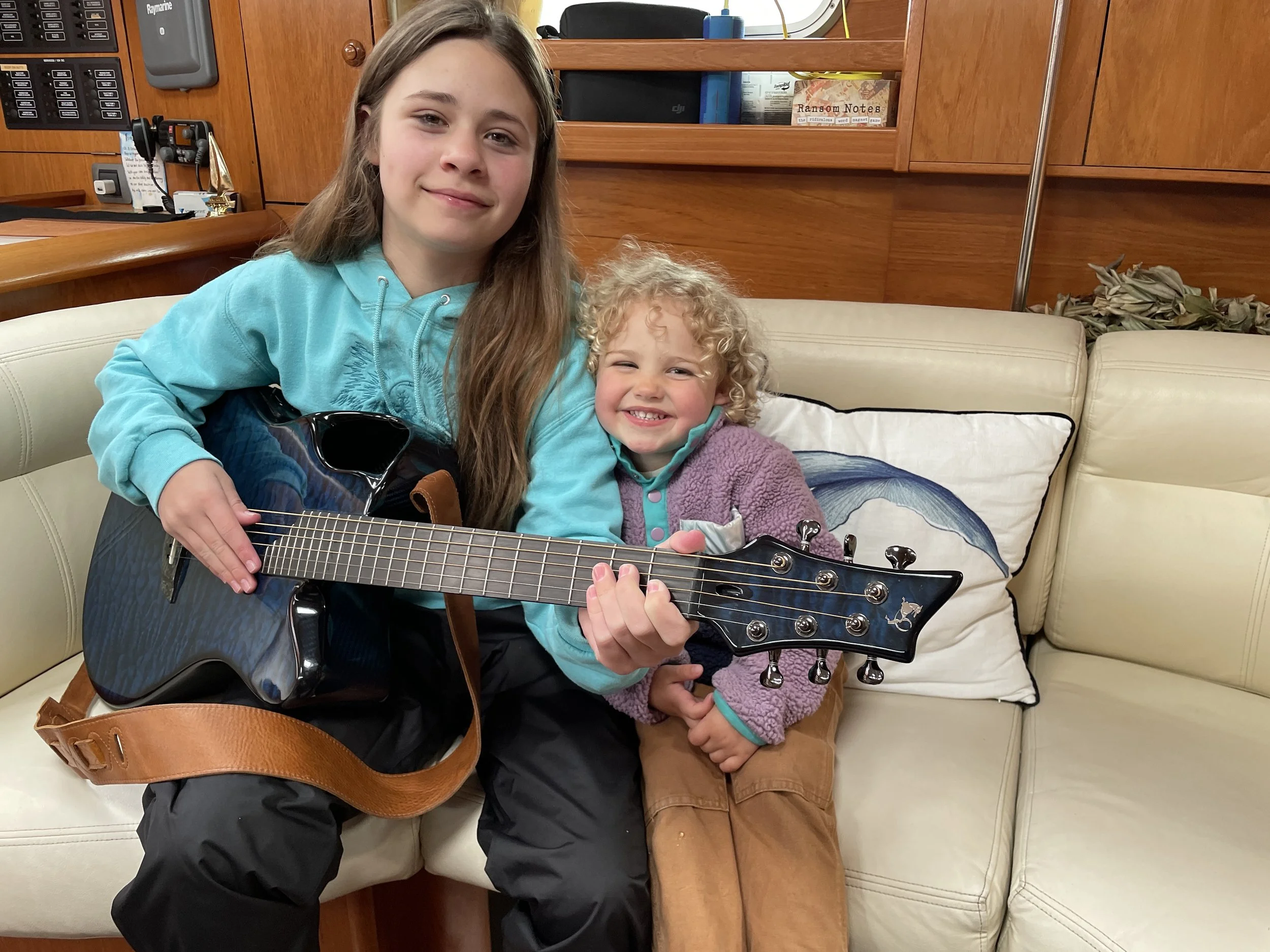 Two young girls sit on a cream-colored couch, with the older girl holding a dark blue acoustic guitar. The older girl has long brown hair and is wearing a turquoise hoodie and black pants, smiling gently. The younger girl has curly blonde hair, weari