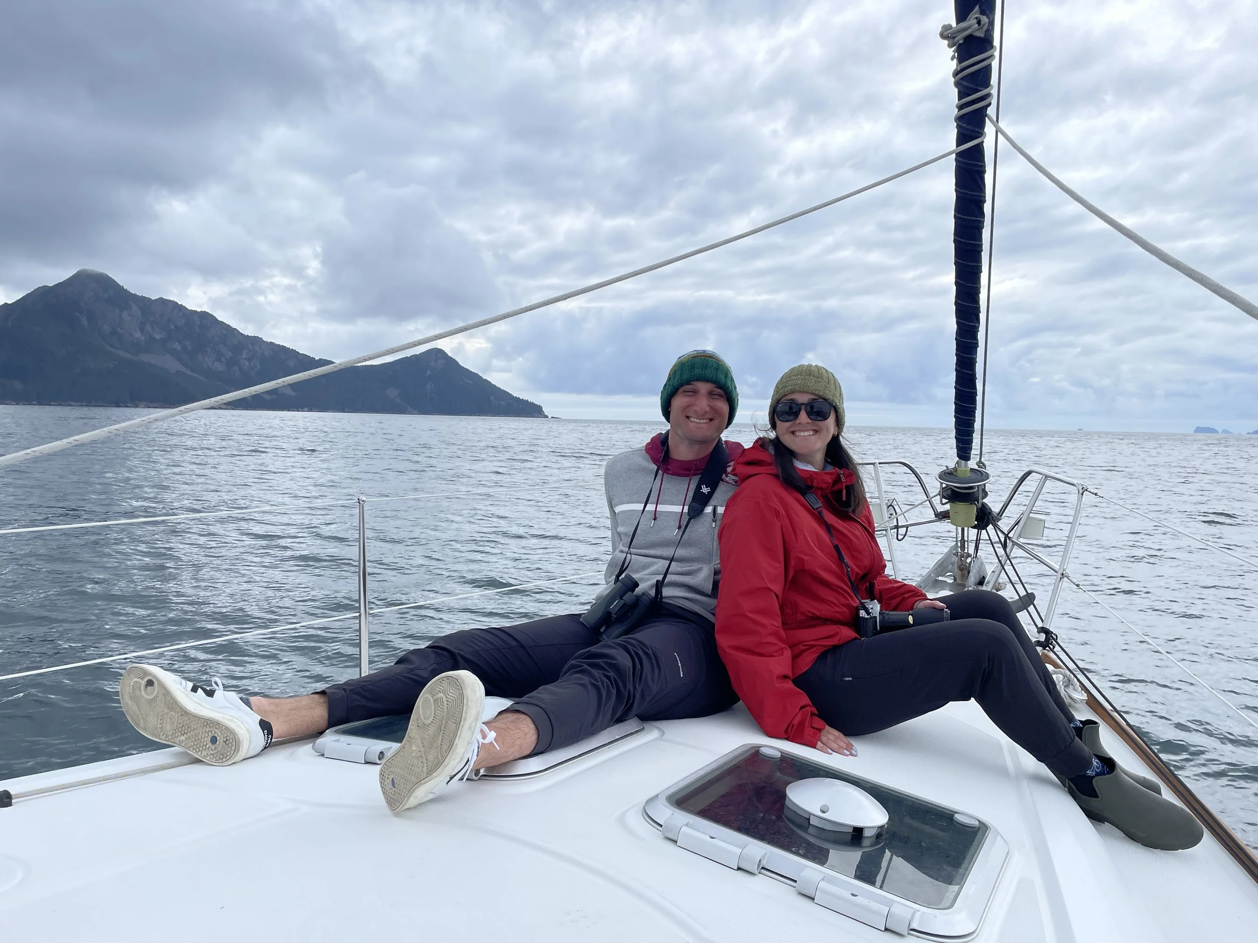 Two people sitting on the deck of a sailboat, smiling, with a mountain and cloudy sky in the background.