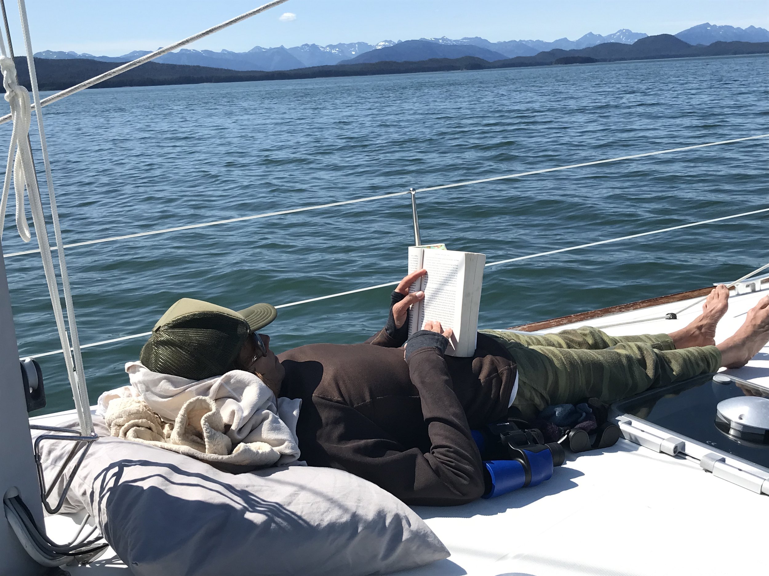 Person relaxing on a boat, reading a book, with mountains and ocean in the background.