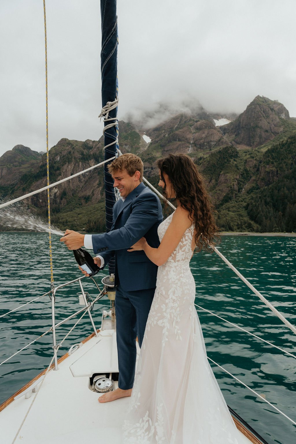 A couple in formal attire celebrates on a sailboat, with a man opening a bottle of champagne and a woman in a white dress in front of mountain scenery.