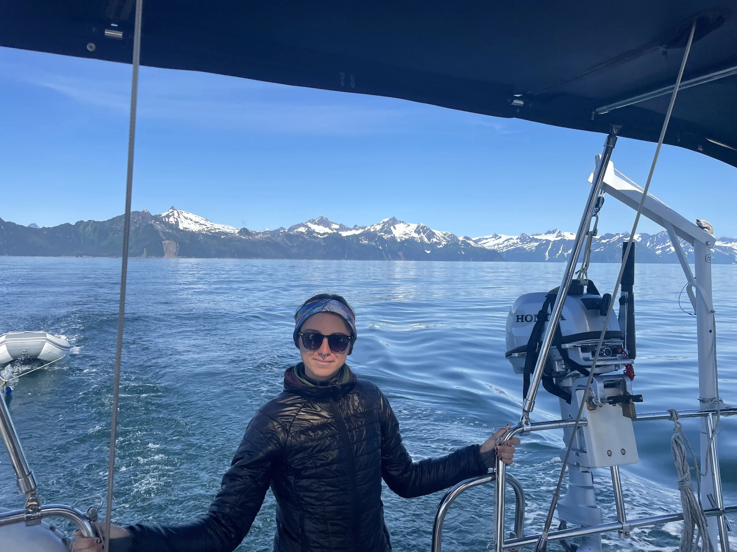 Person wearing sunglasses, a jacket, and a headband, standing on a boat with mountains and snow-capped peaks in the background, holding onto the boat's railing.